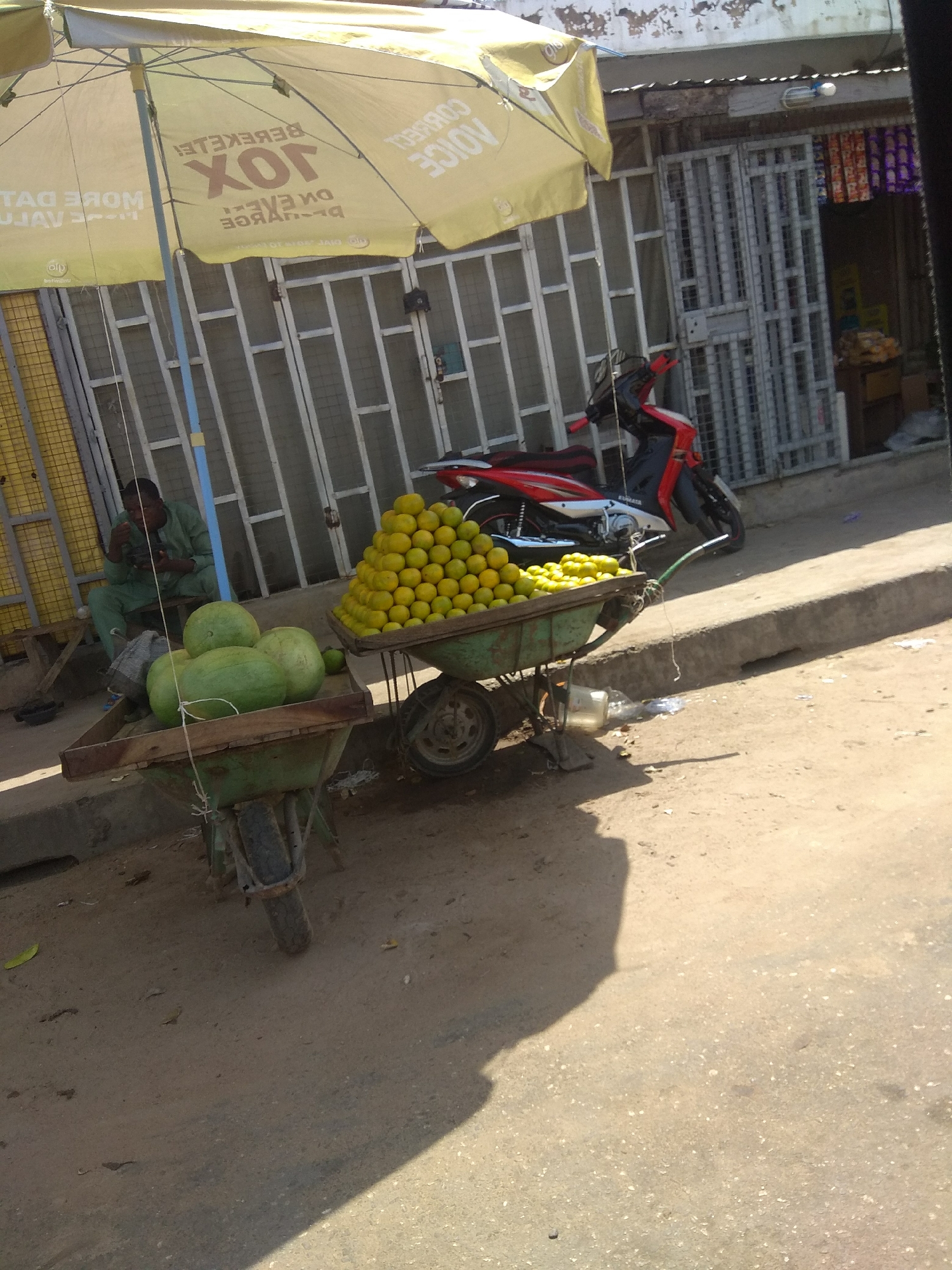 This is a medium shot, eye-level, outdoor photograph taken in Jalingo, Nigeria. In the foreground, a wheelbarrow filled with large green fruits, possibly watermelons, is positioned on a dirt path. To the right, another wheelbarrow is piled high with yellow fruits, likely oranges or lemons. A red and silver motorcycle is parked behind the fruit wheelbarrow. In the background, a man wearing a green traditional outfit is seated on a bench under a large yellow umbrella, eating. Behind him is a storefront with metal bars, and further back, colorful items are visible hanging inside the shop. The scene is lit by natural sunlight, casting shadows on the ground.