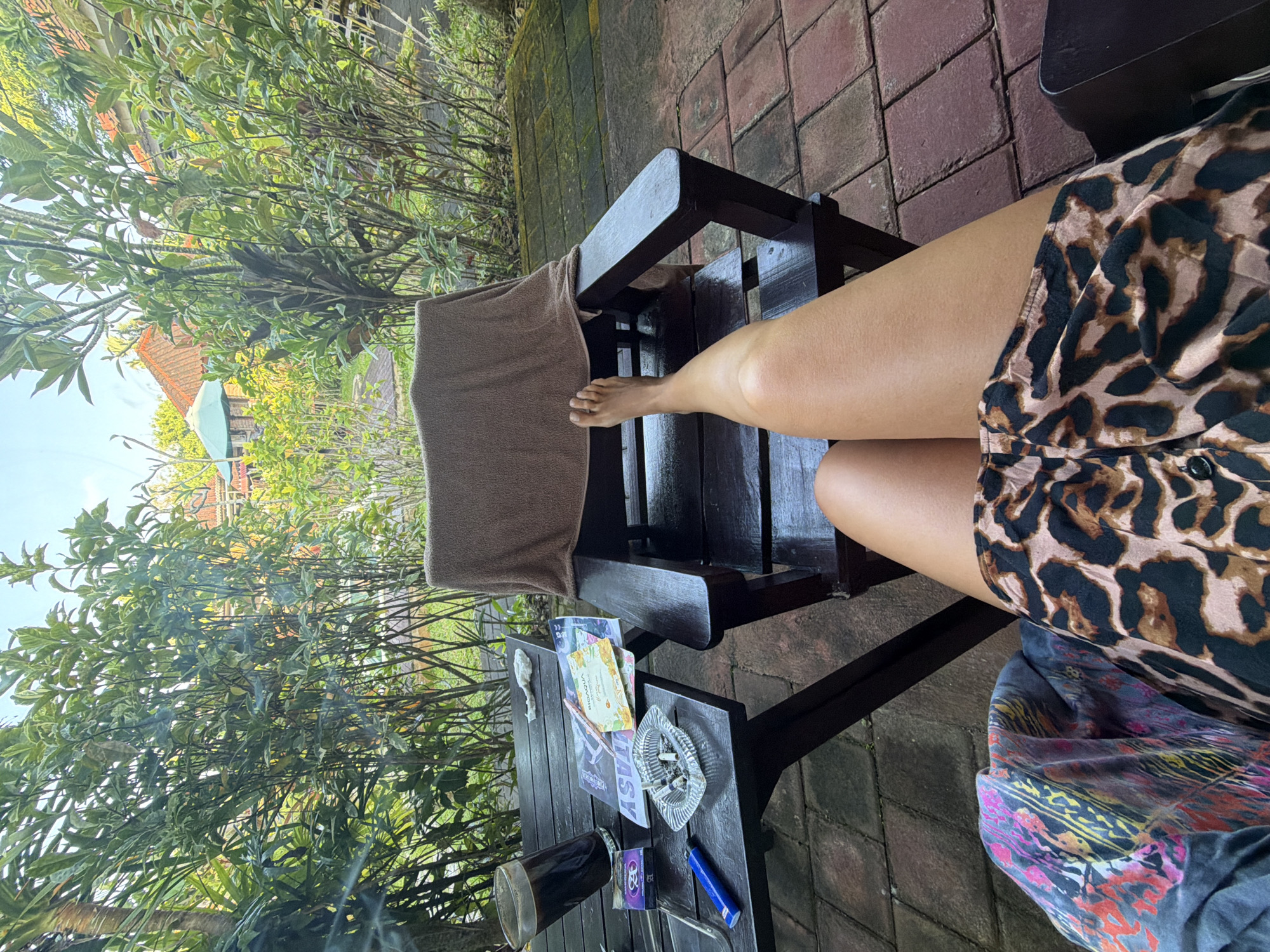 The image depicts a person's legs and part of their leopard-print shorts as they sit on a dark wooden chair outdoors in Kutuh, Indonesia. A brown towel is draped over the chair's back. The person's left foot rests on the chair's slats. A small wooden table beside the chair holds a glass of dark liquid, an ashtray with cigarette butts, a pack of cigarettes, a lighter, and leaflets. Lush greenery is visible in the background, along with a building with a red tiled roof and a blue structure. The ground is paved with red bricks.
