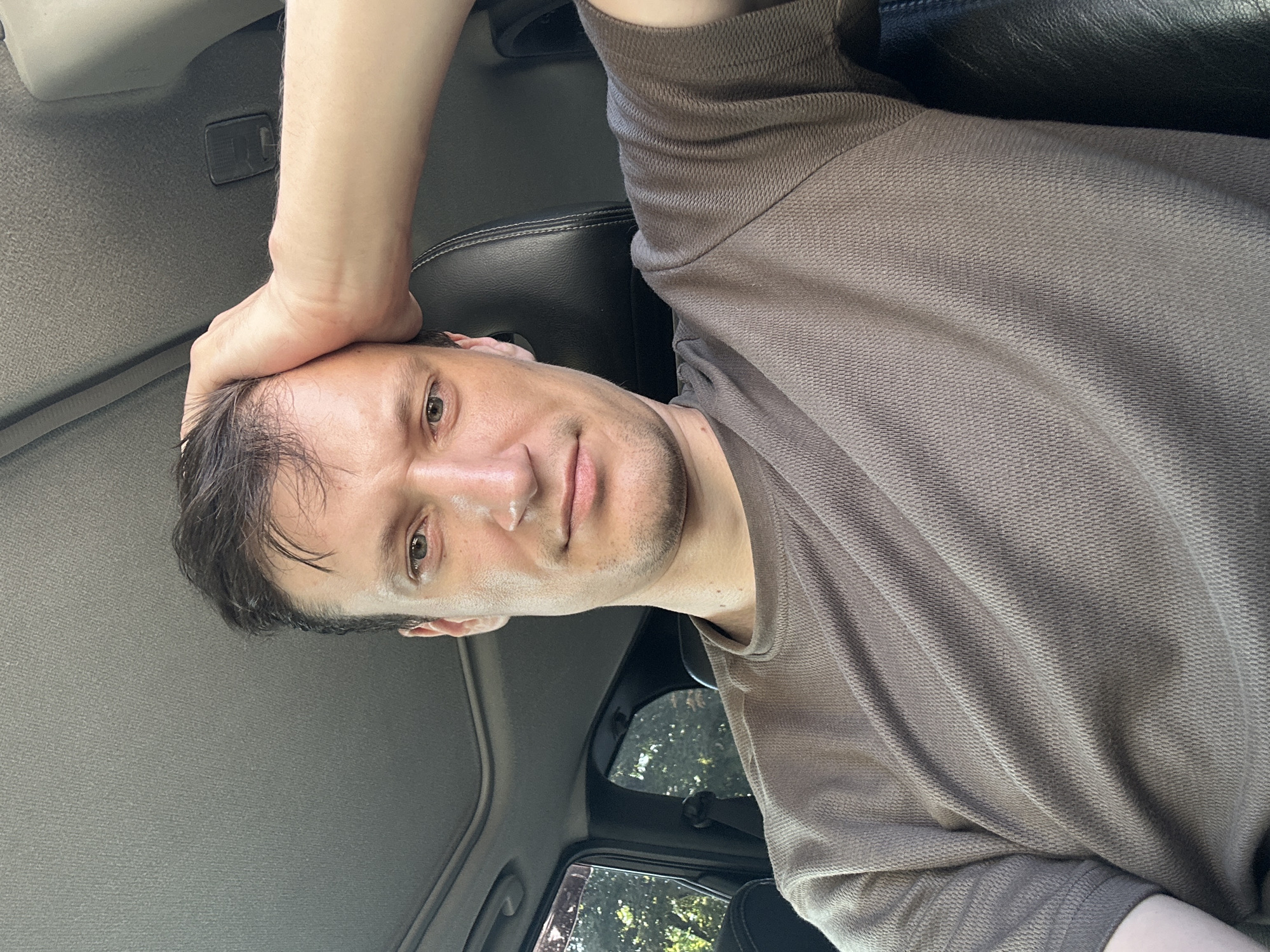 A man is sitting in the driver's seat of a car, with his right arm raised and hand resting on his head. He is wearing a brown t-shirt. The interior of the car is visible, including the ceiling, a sunroof, a dark leather seat, and a window with trees visible outside. The scene appears to be a personal selfie taken during the daytime. There is no information in the image that would indicate a specific location in the city of Kamianets-Podilskyi, Ukraine.