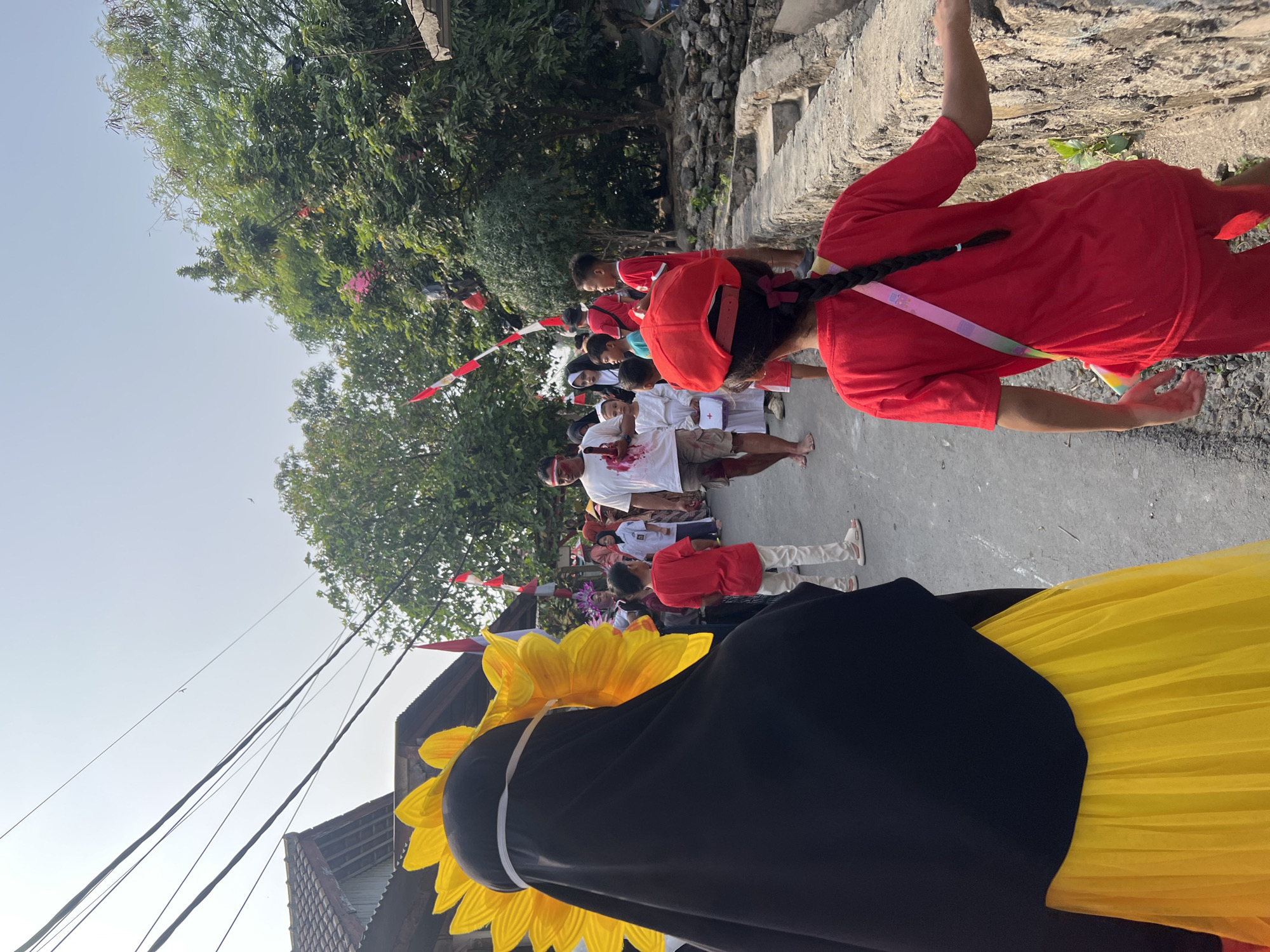 In Klapanunggal, Indonesia, a procession is underway along a paved street lined with homes and trees. A person in the foreground, facing away from the camera, wears a black hijab adorned with a large yellow sunflower headpiece and a yellow tulle skirt. Behind them, a group of children in red shirts and some adults are walking down the street. One adult male wears a white t-shirt with red stains, and a red band around his forehead. Several red and white Indonesian flags are visible, strung across the street. The procession is heading towards the right side of the frame, where more people and trees can be seen. A stone wall runs along the right side of the street.