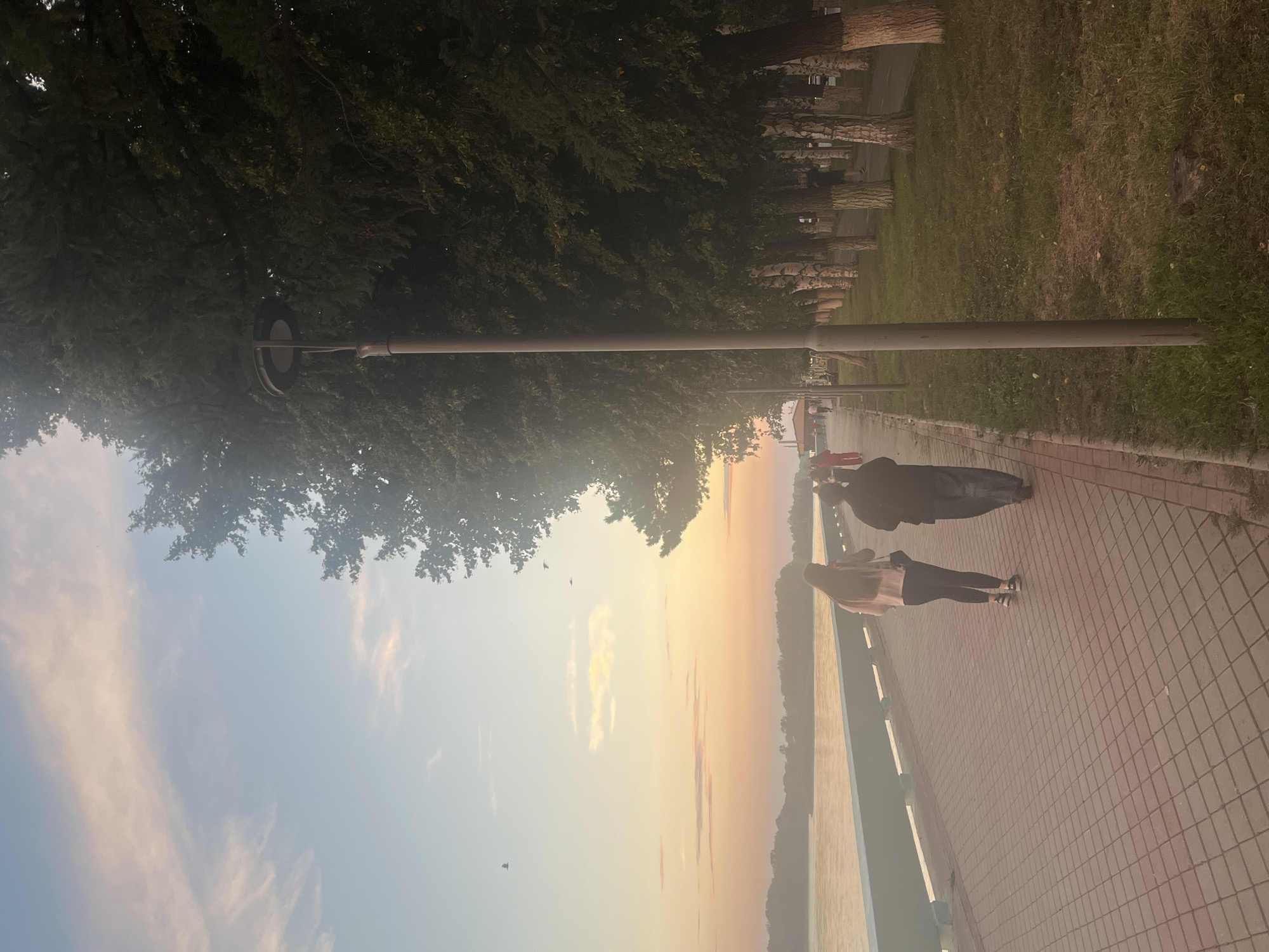 The image depicts a serene sunset scene along a waterfront promenade, likely in Oskemen, Kazakhstan. Two individuals, a woman with long dark hair wearing a light-colored jacket and black leggings, and a man in a dark jacket and grey pants, are walking away from the camera on a paved walkway. The walkway is constructed of red-toned square pavers and is bordered by a railing overlooking a body of water.
To the right of the walkway, a row of trees with dense green foliage forms a natural barrier, interspersed with streetlights. The trees appear to be a mix of species, with some exhibiting white bark. The ground beside the trees is covered in grass. In the distance, more people can be seen strolling along the promenade, and a few birds are visible in the sky.
The sky is a gradient of soft blues and oranges, indicative of a sunset, with wispy clouds adding to the picturesque atmosphere. The water in the foreground reflects the warm hues of the sky. The overall impression is one of tranquility and a leisurely evening stroll by the water.