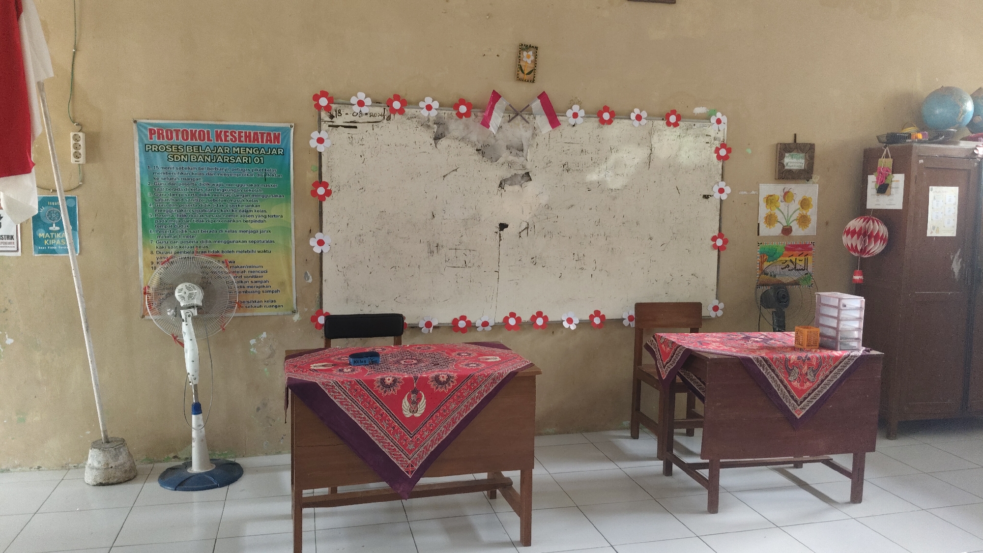 The image depicts a classroom in SDN Banjarsari 01, located in the city of Banjarsari, Indonesia. The room has tiled floors and beige walls, with a large, worn-out whiteboard dominating the center of the back wall. The whiteboard is decorated with red and white paper flowers and two small Indonesian flags. To the left of the whiteboard, a poster titled "PROTOKOL KESEHATAN PROSES BELAJAR MENGAJAR SDN BANJARSARI 01" is displayed, outlining health protocols for learning. A standing fan is positioned in front of this poster.
Two wooden desks are visible in the foreground, each covered with a decorative red cloth. The desk on the left has a blue box on it, and the desk on the right has a small plastic organizer with drawers and an orange basket. A wooden chair is placed behind each desk.
On the right side of the image, a large, dark wooden cabinet stands against the wall. Above the cabinet, a globe and another smaller spherical object are placed. A decorative red and white lantern hangs from the cabinet. Various pictures and artwork are also displayed on the wall, including a framed picture of sunflowers and another with Arabic calligraphy. A white pole with a circular concrete base stands near the left edge of the image. The overall scene suggests a functional classroom environment, likely for elementary school students, prepared with safety protocols and decorated for a celebratory or educational purpose.