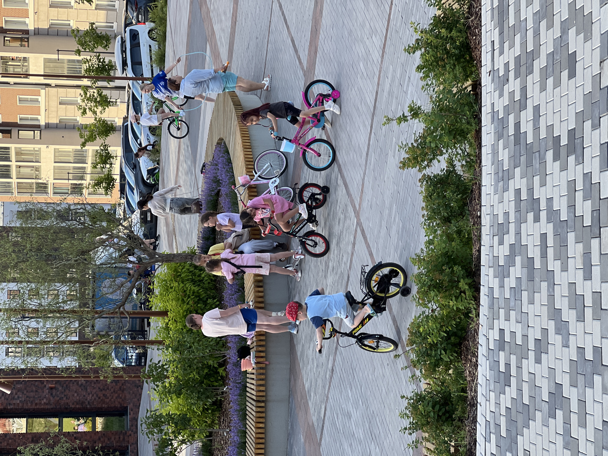 In a paved outdoor area with a patterned brick walkway, several children are engaged in various activities. The scene is set during the daytime, with bright sunlight casting shadows, suggesting fair weather. A group of children are gathered, some on bicycles, others standing or sitting.
One child, wearing a blue t-shirt and a red cap, is on a black bicycle with yellow accents and training wheels, appearing to be practicing riding. Another child, in a dark t-shirt and shorts, is on a pink bicycle. Further down, a child in a pink top and shorts is also near a bicycle. A young person in a light blue shirt and white shorts is on a green bicycle, holding a hula hoop. Two other individuals, one in a white shirt and light blue shorts and another in a grey t-shirt and dark shorts, are also present among the children.
In the background, there are multi-story residential buildings with many windows and some greenery, including trees and bushes. The overall atmosphere appears to be one of casual outdoor recreation and play. No visible text is apparent in the image.