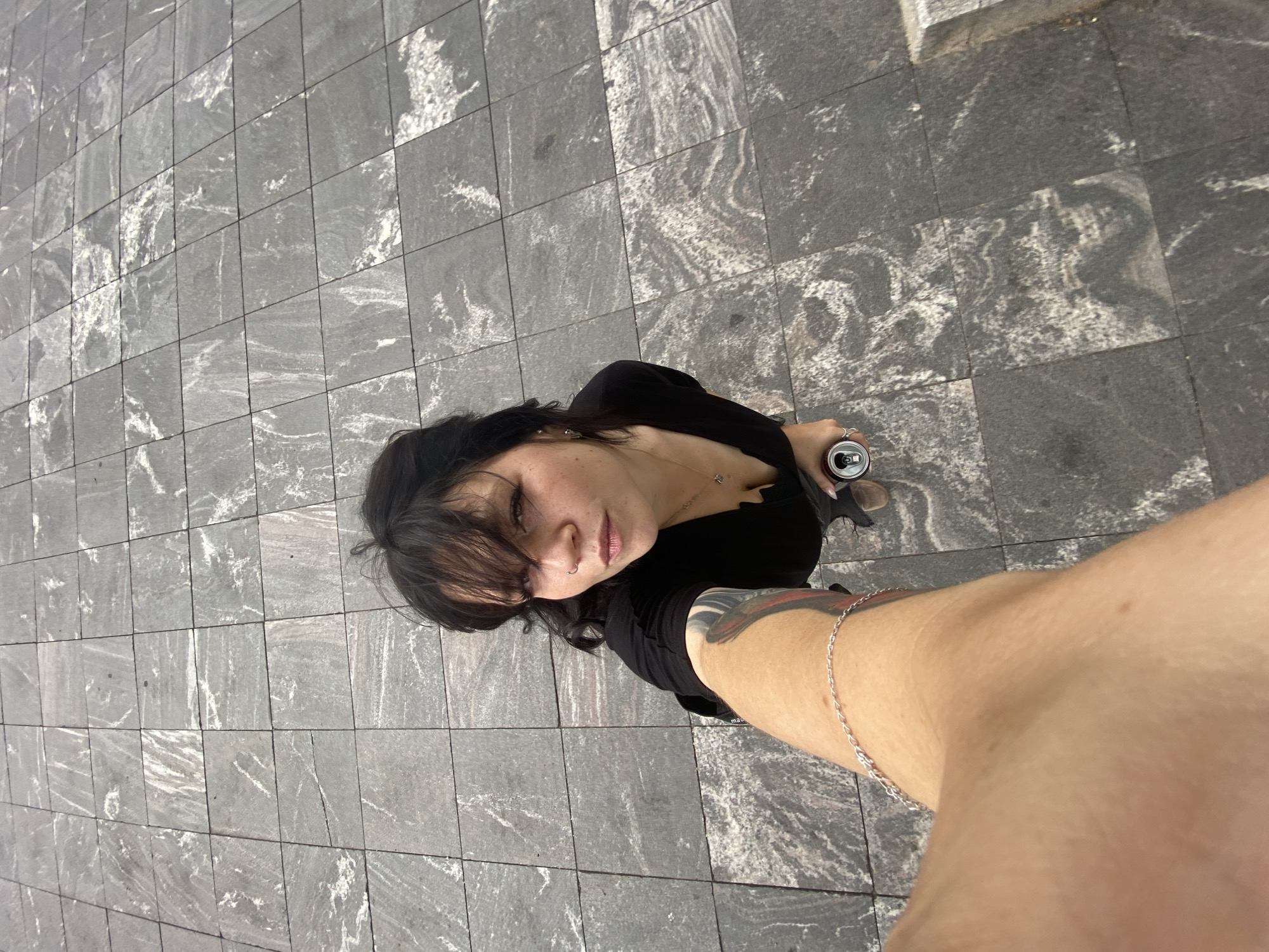 An overhead, wide-angle selfie taken from a first-person perspective shows a person holding a soda can and the camera. The subject, a woman with dark hair and bangs, wears a black V-neck top. She has a tattoo on her left arm and is wearing a silver bracelet and necklace. She is standing on a paved surface made of large, grey square tiles with white marbling. The background is out of focus, but appears to be a city street with some structures visible. The lighting suggests it is daytime. The image was taken on Krestovsky Island in Saint Petersburg, Russia.