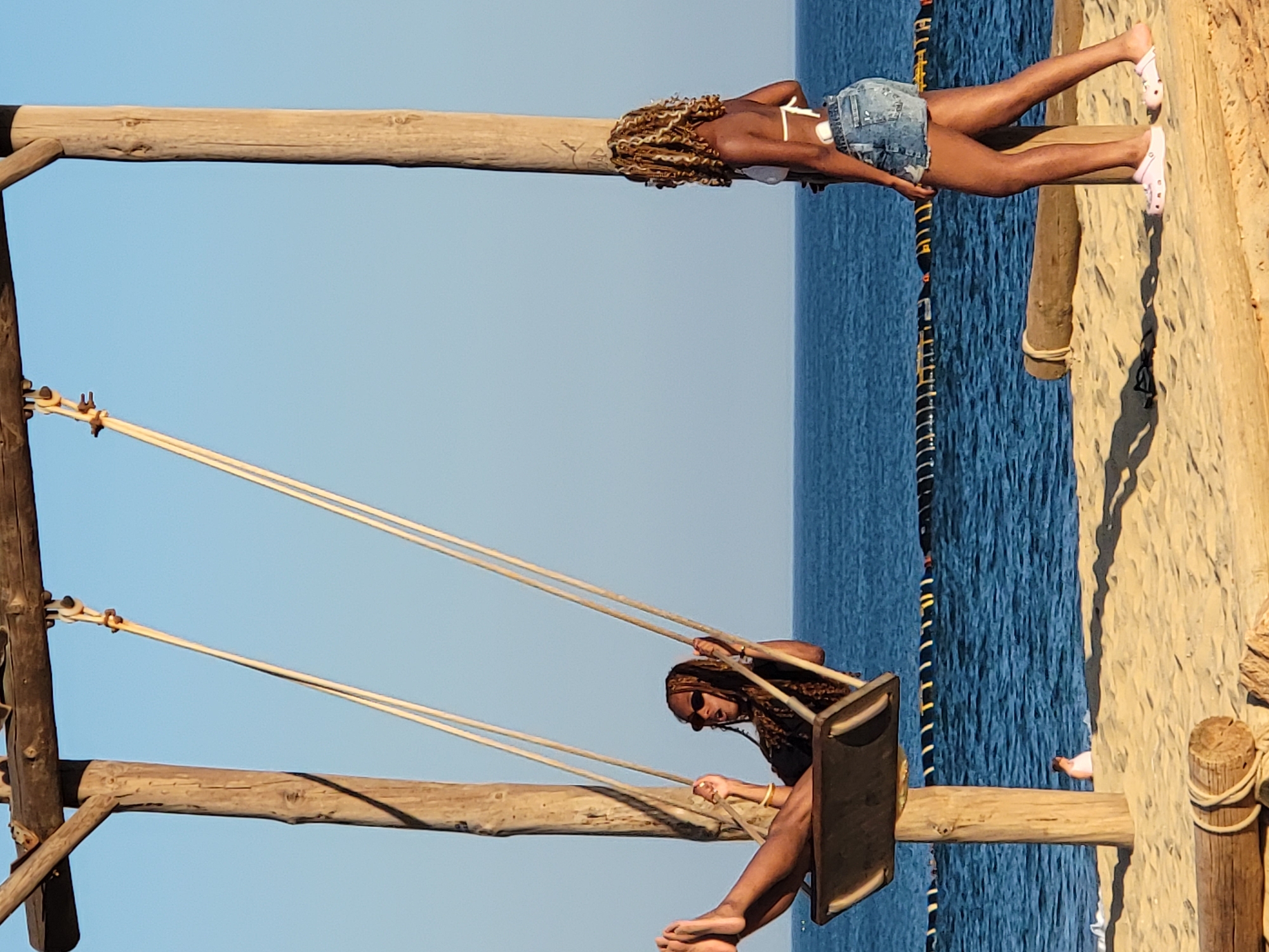 A young woman with braided hair and sunglasses is on a wooden swing suspended by ropes from a wooden frame. She is wearing dark clothing and has her legs extended. She is looking towards the viewer and her mouth is open. Another young woman with blonde braided hair is standing to the right of the swing, facing away from the viewer. She is wearing a white bikini top and denim shorts. She is looking towards the ocean. The swing and the standing woman are on a sandy beach. In the background, the blue ocean stretches to the horizon. A yellow and black floating barrier is visible in the water. The scene appears to be during the daytime, with clear blue skies. The location is the beach in Quarteira, Portugal, a popular tourist destination known for its beaches and coastal promenade.