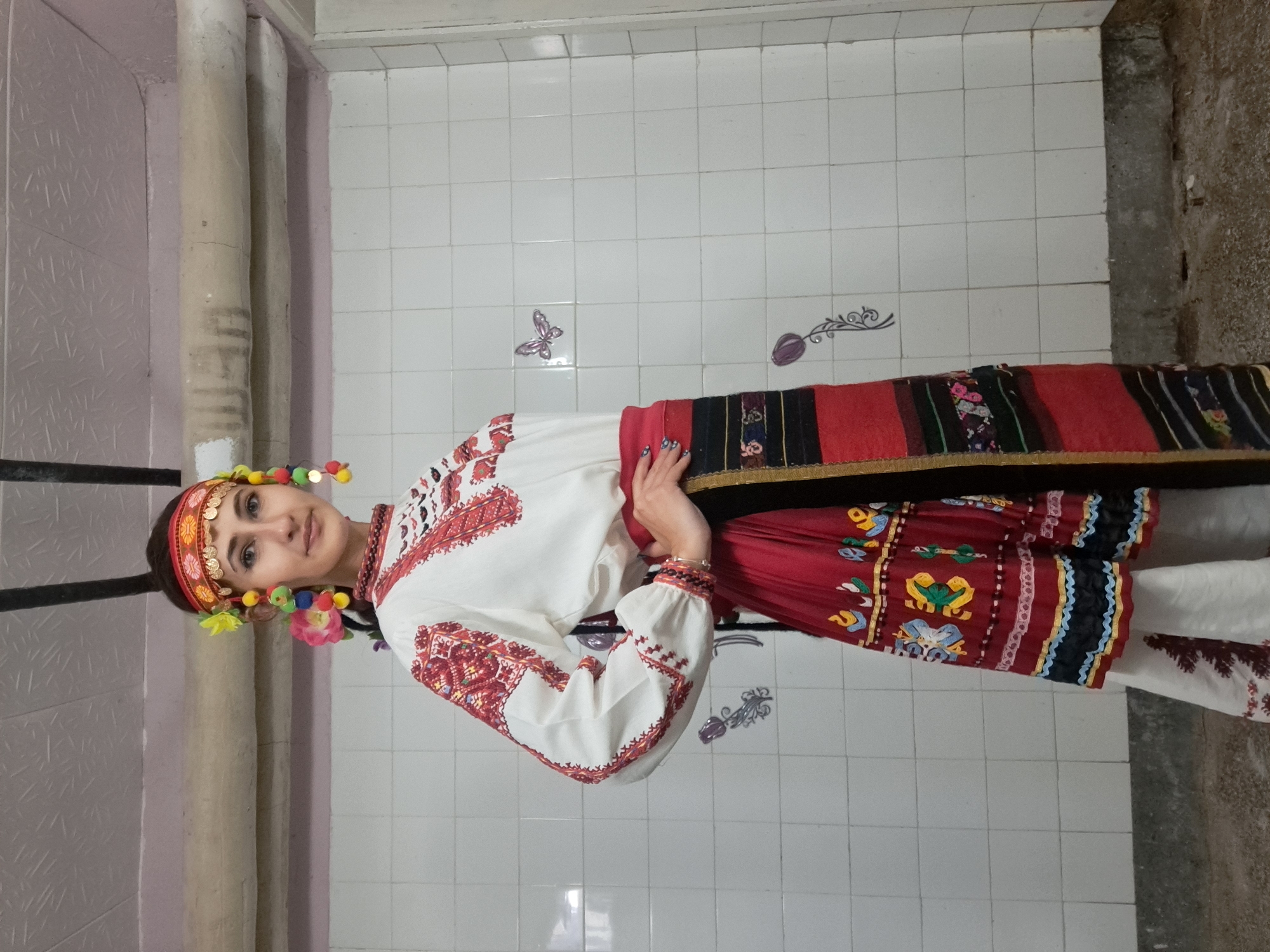 The image shows a young woman dressed in a traditional Bulgarian folk costume. She has a colorful headband decorated with flowers and pom-poms. Her blouse is white with red embroidery on the sleeves and around the neckline. She wears a wide, multicolored woven belt and a red skirt with intricate patterns. The background is a tiled wall and a corner with a concrete wall. The image was taken in Lokorsko, Bulgaria.