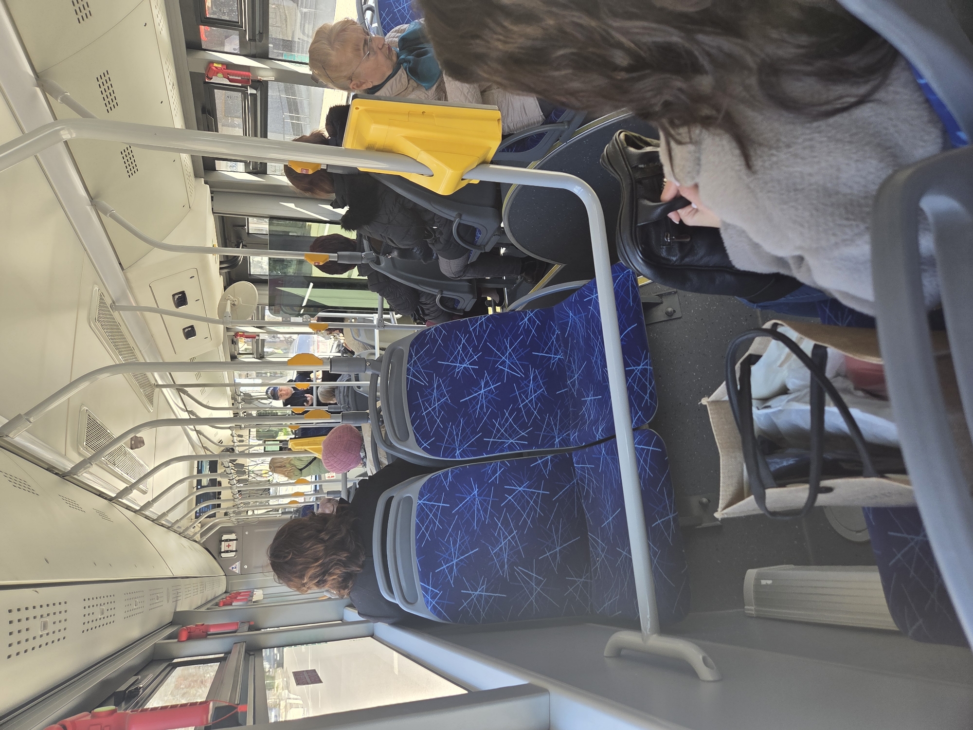 The image shows the interior of a public bus in Pleven, Bulgaria. The bus is occupied by several passengers, including an elderly woman wearing glasses and a light-colored jacket, and individuals in winter attire such as coats and hats. The bus features blue patterned seats, yellow hand grips, and overhead handrails. A black leather bag is visible at the feet of a passenger in the foreground. The image is taken from the perspective of someone sitting on the bus, looking towards the front. The interior of the bus is well-lit, likely due to daylight coming in through the windows. The windows appear to be partially open. There are red emergency hammers visible attached to the interior walls of the bus.