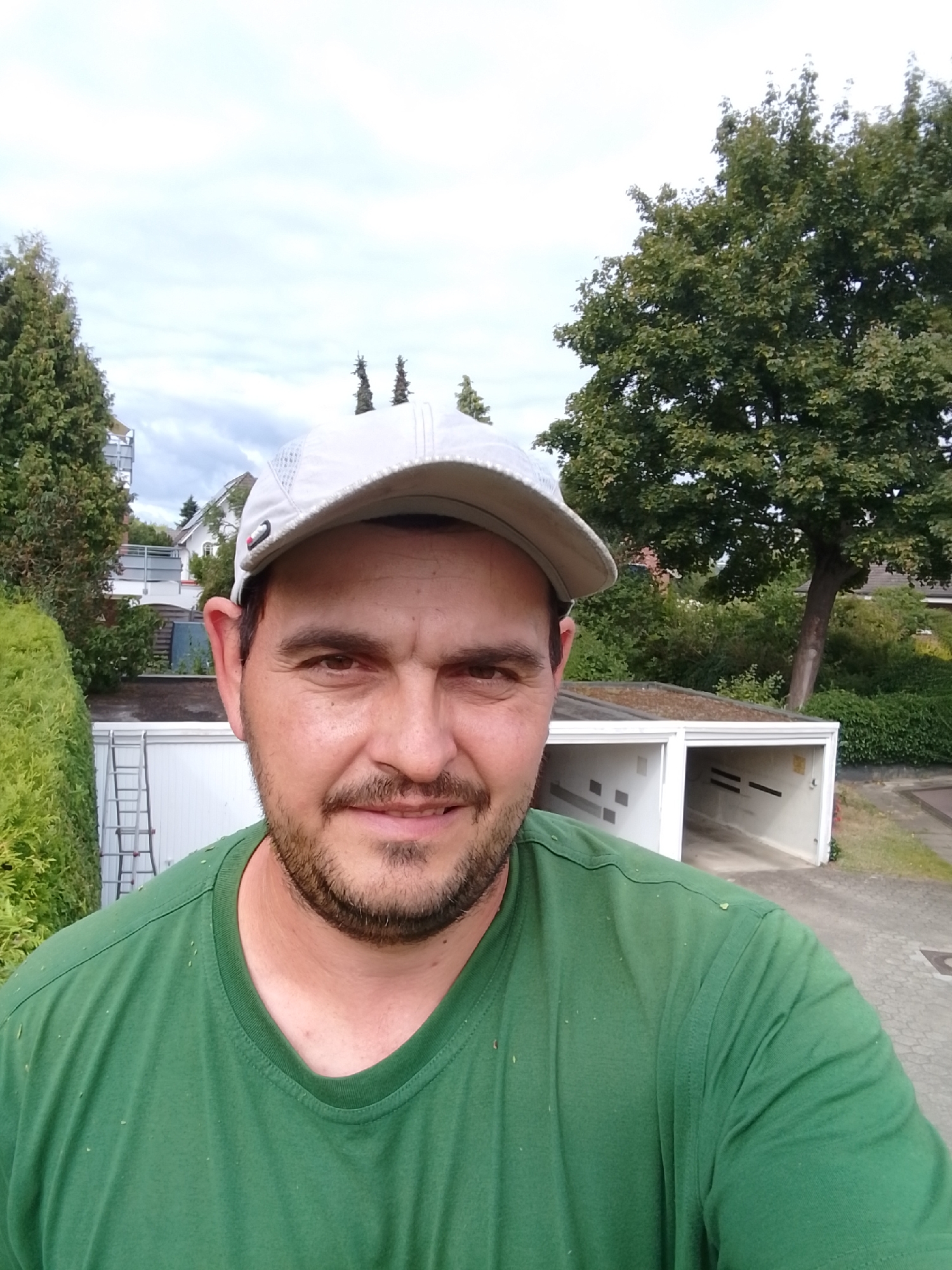 A man wearing a green t-shirt and a grey baseball cap takes a selfie outdoors. He has brown hair, a beard, and is smiling. Behind him, a large green tree is visible, along with a white garage and some bushes. The location is Wedel, Germany.