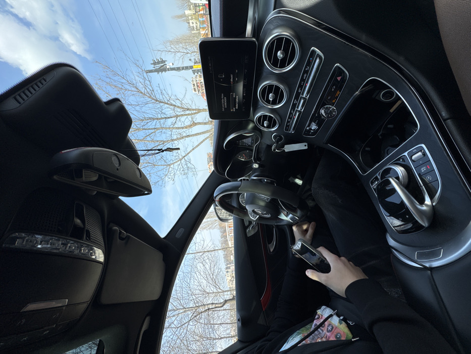 The image depicts the interior of a black Mercedes-Benz vehicle, viewed from the front passenger side, looking towards the driver and dashboard. A person, identified as the driver, is seated, wearing a black long-sleeved shirt and black trousers, holding a silver-colored smartphone (appearing to be an iPhone) in their left hand. The vehicle appears stationary. The steering wheel, partially obscured by the driver's arm, is a three-spoke design with a black rim and a red accent line at the 12 o'clock position. Car keys on a white lanyard are visible in the ignition slot to the right of the steering wheel.
The infotainment screen on the dashboard displays information in German, showing "Zart und Zartgenuss" and "Musik" (Music) along with "iPhone" as a connected device, and the time "09:00". The ambient temperature is indicated as "18.0". The center console features a prominent silver control knob and touchpad, surrounded by various buttons. Three circular air vents are visible above the climate controls. An overhead console contains interior lighting and control buttons, with a sun visor in the raised position. A small, dark ornament hangs from the rearview mirror.
Through the front windshield and side windows, an urban exterior scene is visible under a clear blue sky with scattered white clouds. The environment features numerous bare deciduous trees, indicating a late winter or early spring season. In the background, modern multi-story buildings are present, with one building on the right displaying sections of colorful graffiti. A black traffic light pole with traffic signals is visible on the right side of the road, and overhead power lines are discernible against the sky. The scene is located in an urban area of Sofia, Bulgaria.