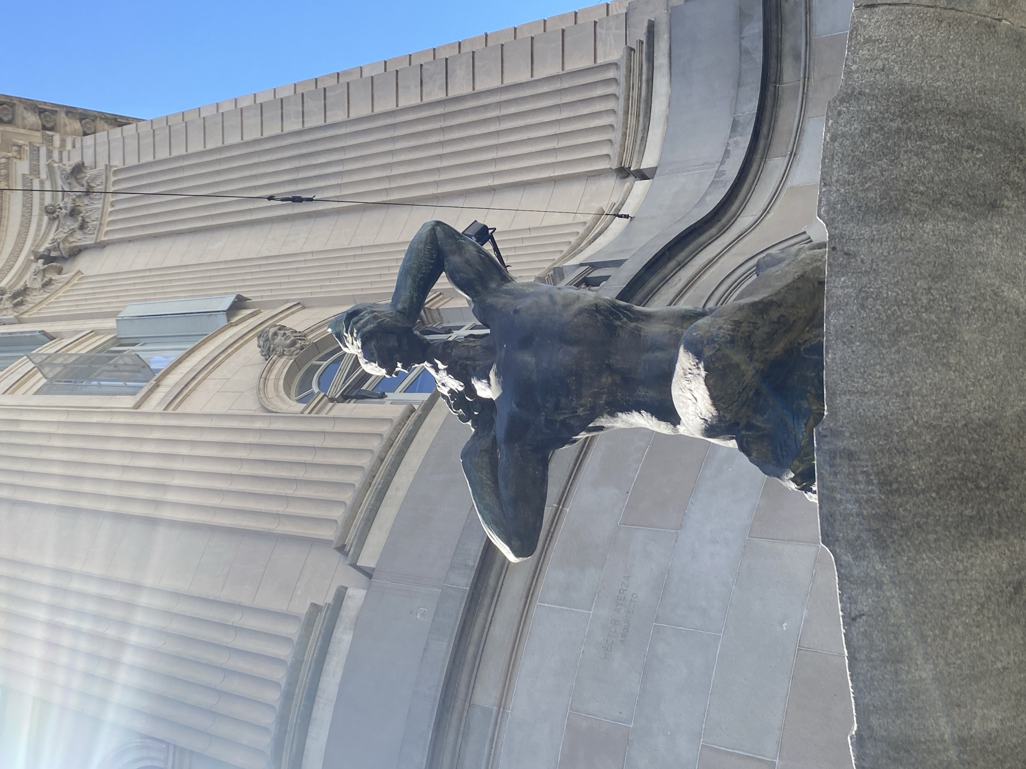 The image shows a bronze sculpture of a man, seemingly deep in thought, set against the backdrop of a building facade in Buenos Aires, Argentina. The building features Art Deco architectural details with horizontal lines and arched windows. The name "HECTOR AYERZA, ARQUITECTO" is etched on the building. Overhead power lines cross in front of the facade. The sun shines from the left, creating lens flare. The sculpture is positioned on the corner of the building.