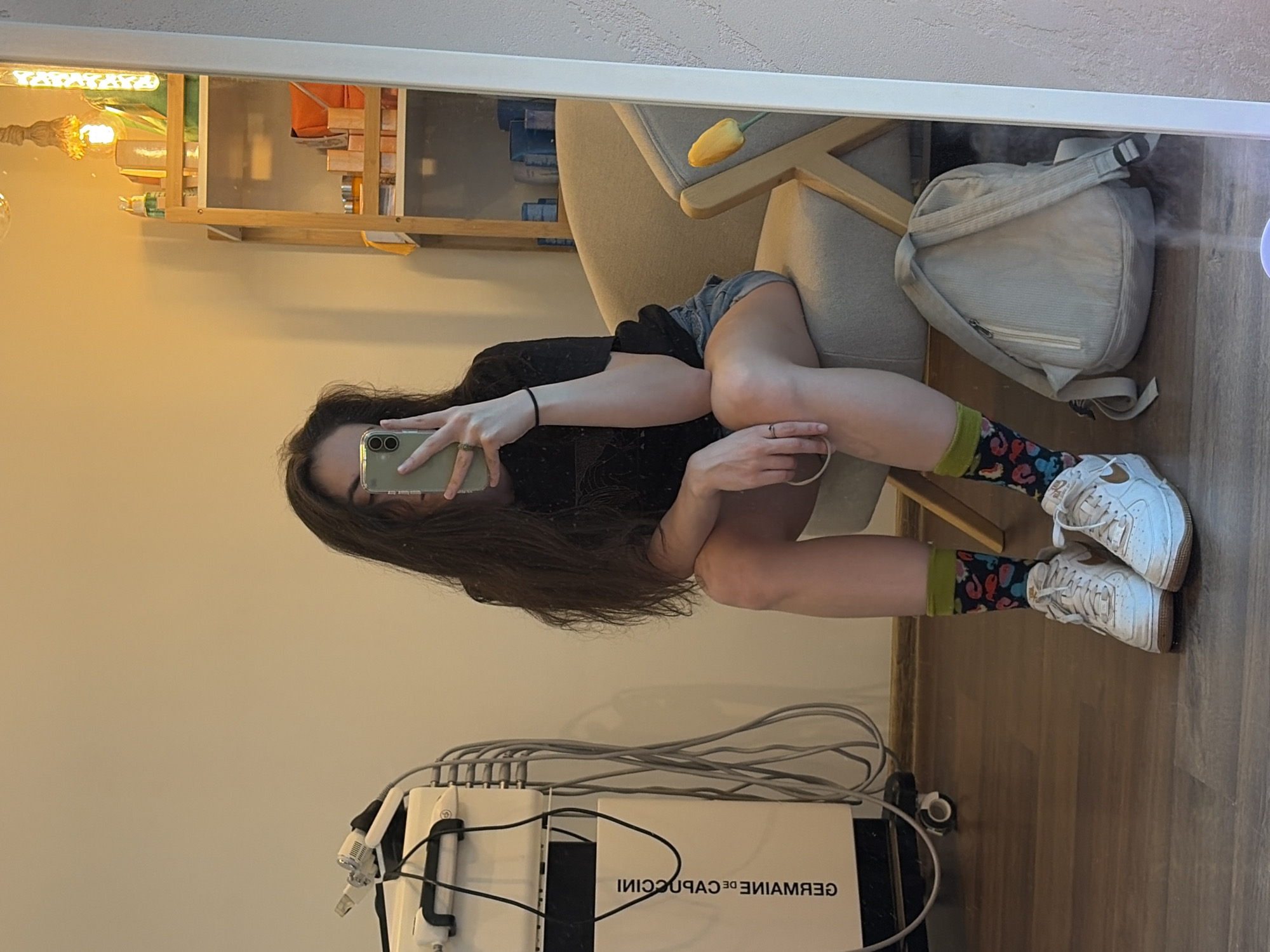 A person is taking a mirror selfie in what appears to be a room. The person is sitting down, leaning back on their hands. They are wearing a black t-shirt, denim shorts, colorful patterned socks, and white sneakers. Their long brown hair is falling over their shoulders. In the reflection, a light-colored chair with a yellow tulip on it is visible, along with a shelf filled with various items and a beige backpack. A medical-looking device with numerous cables is present in the foreground of the room. The flooring is wood. The lighting suggests it is daytime. Visible text on the device reads "GERMAINE DE CAPUCCINI".