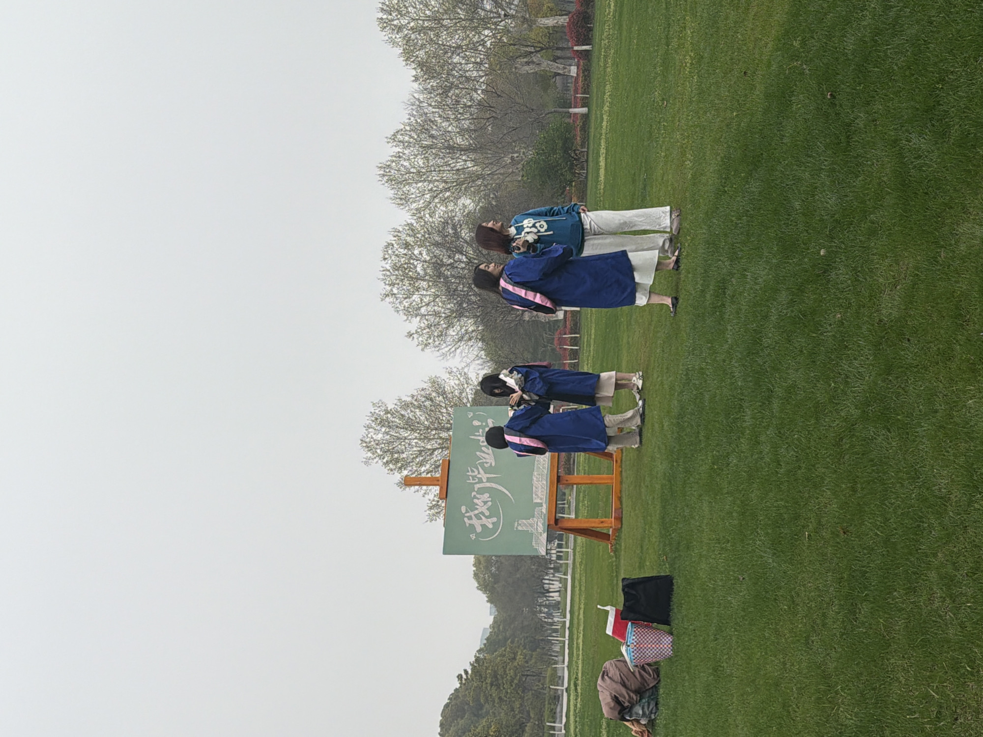 The image shows a group of people on a grassy lawn in Jiangning District, China. There are four people standing near an easel displaying Chinese characters. Two of the individuals are dressed in what appears to be graduation gowns. A woman is seen in the top right wearing a blue sweater with white pants and another in a blue gown, likely taking pictures. Below the easel, bags and other items are placed on the ground. The environment is characterized by a grassy field with trees in the background and an overcast sky.