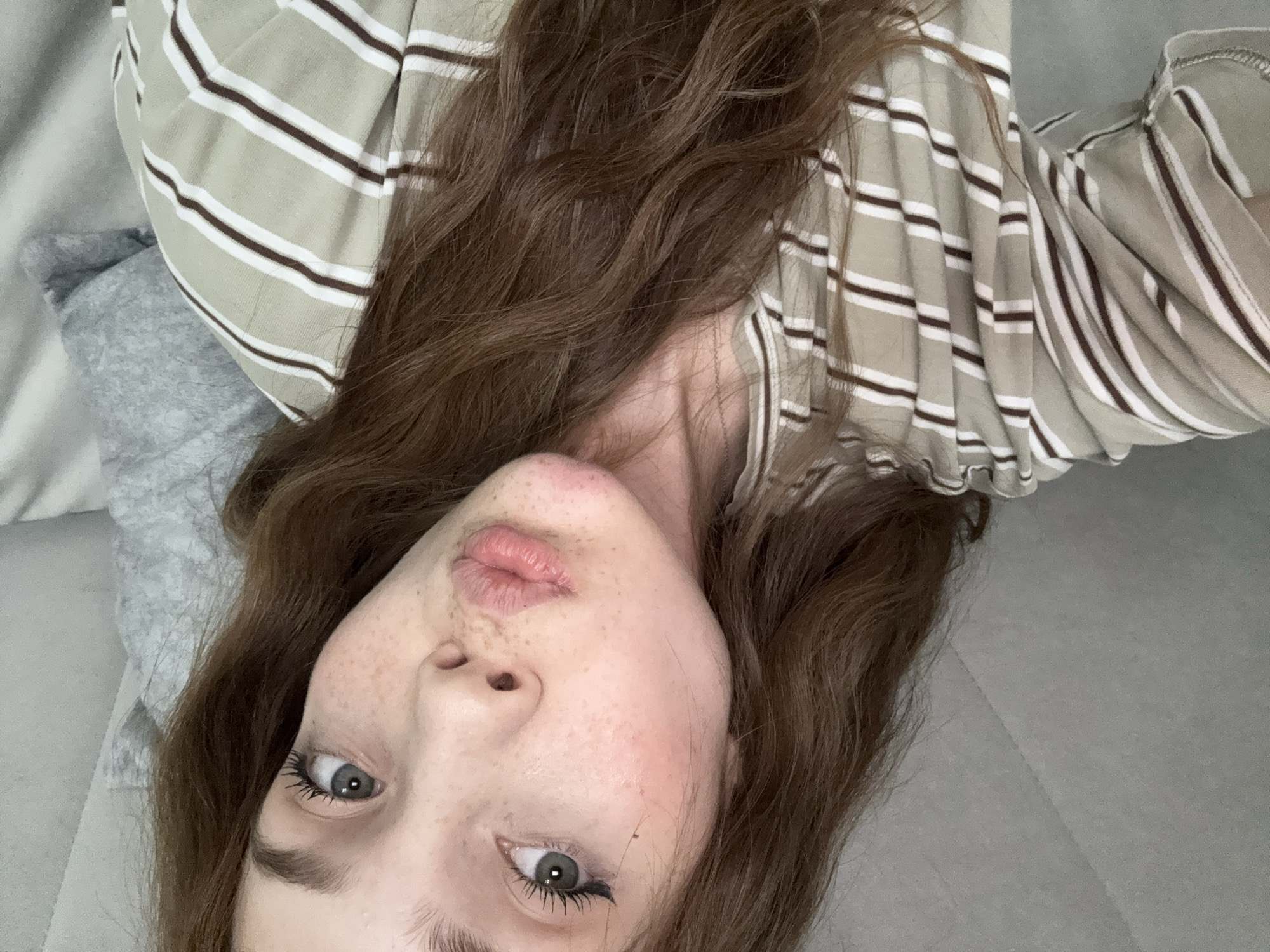 This media file captures a close-up, inverted selfie of a young individual, likely female, with long, wavy, reddish-brown hair. The subject is lying down, looking directly upwards into the camera, resulting in the image being oriented upside down.
**Subjects:** The primary subject is a young person. Their face is visible, characterized by prominent freckles across the nose and cheeks. They have striking green or hazel eyes, with visible mascara applied to the eyelashes. The lips are slightly parted in a soft pout.
**Objects:** The individual is wearing a striped top, featuring alternating bands of beige or light olive green, white, and dark brown stripes. One of the cropped images shows a ruffled edge, possibly on a sleeve or neckline. In the background, a soft, textured grey fabric, resembling a pillow or blanket, is partially visible, suggesting an indoor setting like a bed or couch.
**Scene and Setting:** The scene appears to be an intimate, indoor setting, likely a bedroom or living area. The background elements are simple, consisting of fabric and possibly a plain wall or another surface, in muted grey tones. The overall lighting suggests ambient indoor light.
**Activity or Event Taking Place:** The subject is engaged in taking a selfie, posing for the camera from a reclining position.
**Notable Details:**
* **Orientation:** The image is distinctly inverted, with the subject's head at the bottom of the frame and hair flowing upwards.
* **Facial Features:** The freckles are a prominent detail, as are the subject's expressive eyes and the subtle pout.
* **Hair:** The long, wavy, rich brown hair frames the face and spreads across the background.
* **Clothing Pattern:** The striped pattern of the top is a key visual element, with its distinct color combination.
* **Emotions:** The expression appears casual and slightly playful, perhaps a common selfie pose.
* **Location Cues:** No specific external location cues are present; the setting is generalized to an indoor space.
**Visible Text:** There is no visible text in the image.
