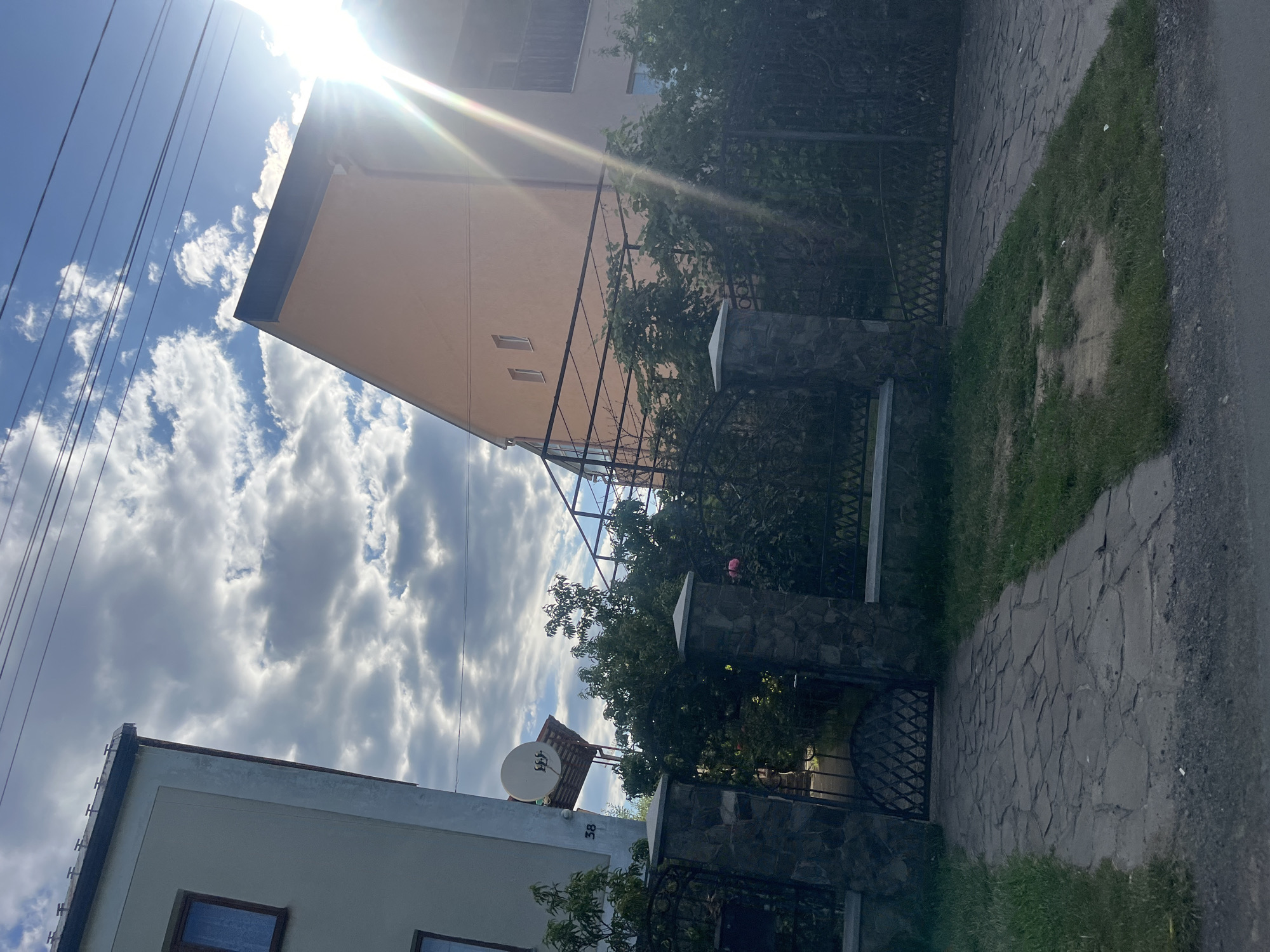 In Storozhnytsia, Ukraine, a residential street scene is captured under a partly cloudy sky with bright sunlight glinting. Two multi-story buildings are visible: a light-colored building on the left with a dark-framed window and a satellite dish mounted on its side, and a peach-colored building on the right with multiple windows and a balcony. A paved pathway made of irregular stone slabs leads towards a wrought-iron gate, flanked by stone pillars topped with concrete caps. Overgrown green bushes and trees obscure parts of the property behind the fence, with a hint of a rose bush visible. Power lines are strung across the top of the image. The scene depicts a typical residential setting with elements of architecture and landscaping.
