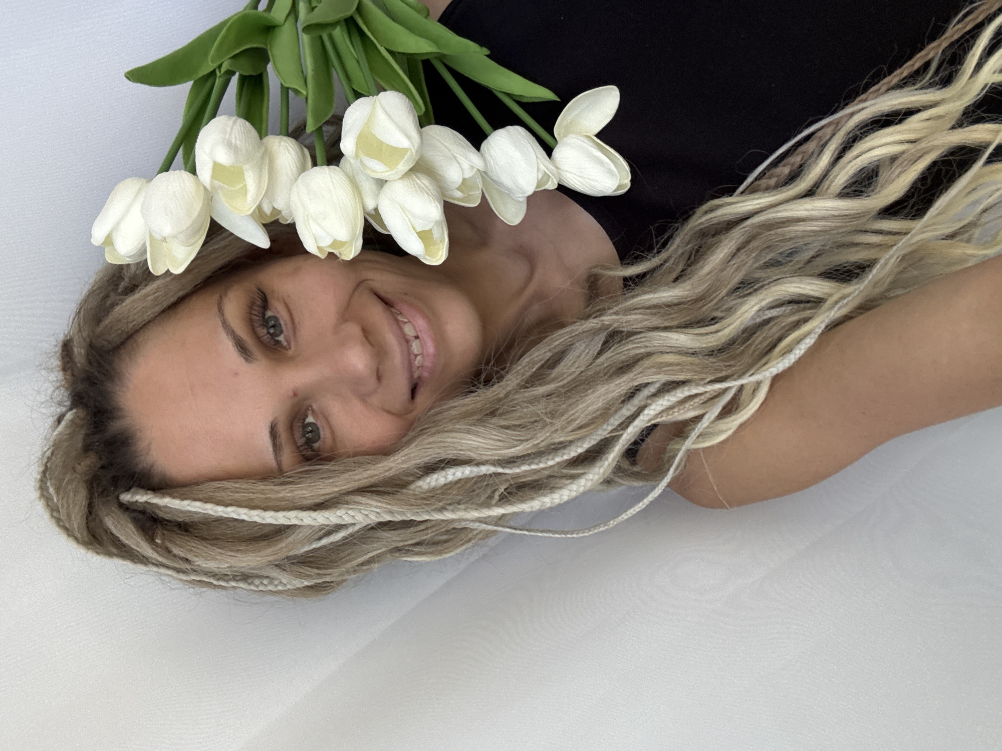 A woman with braided blonde hair is lying down, smiling at the camera. She is wearing a black top and has a bouquet of white tulips near her head. The background appears to be a white surface. The location in the city of Rusanovo, Russia cannot be determined from the image.