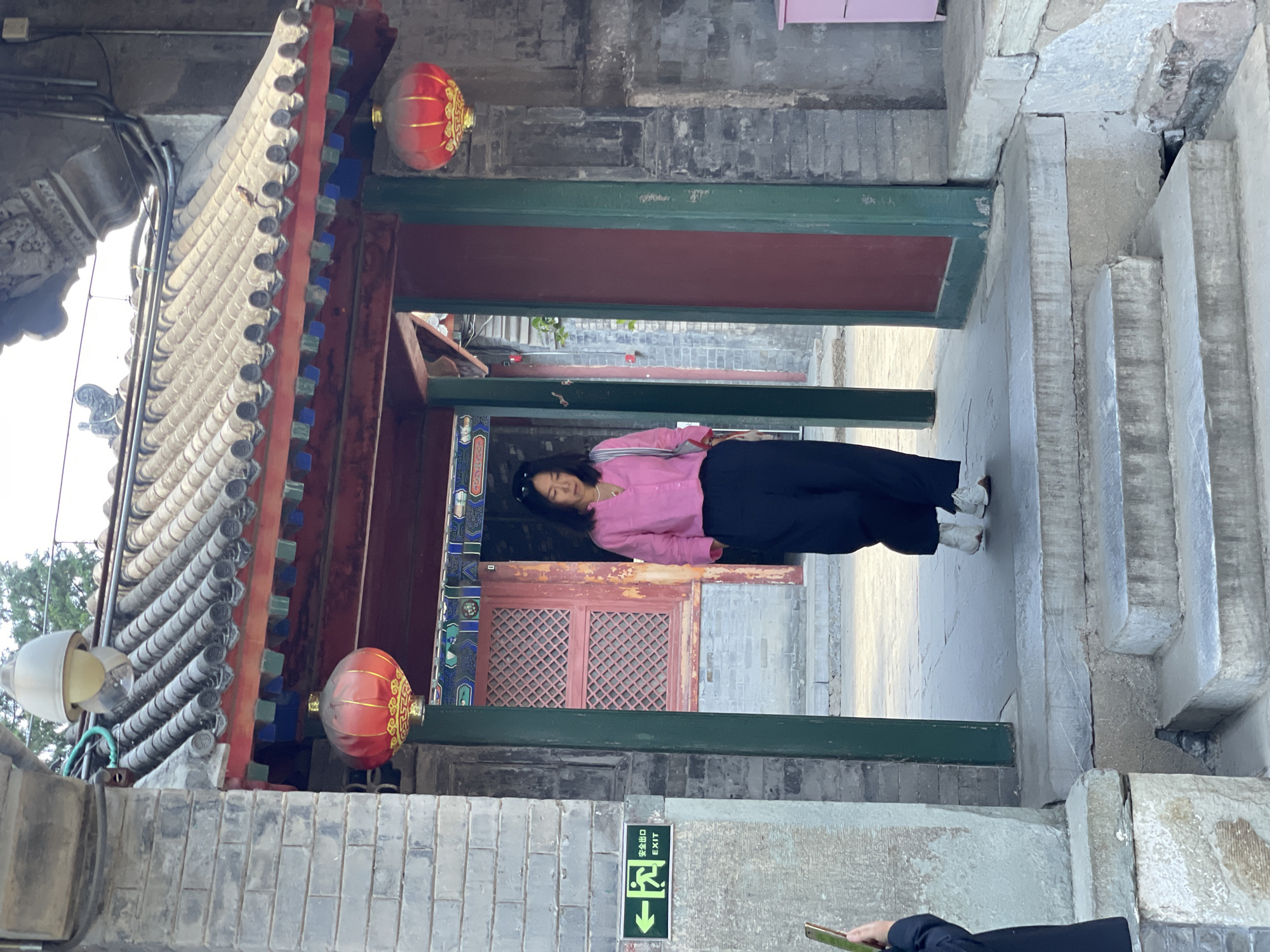A woman in a pink shirt and black pants stands in a walkway under traditional Chinese architecture. The walkway is framed by stone and brick structures with decorative elements, including red lanterns. An exit sign is visible on a wall. The image appears to have been taken in the Xicheng District of China, based on the architectural style. A person holding a smartphone is partially visible in the foreground.