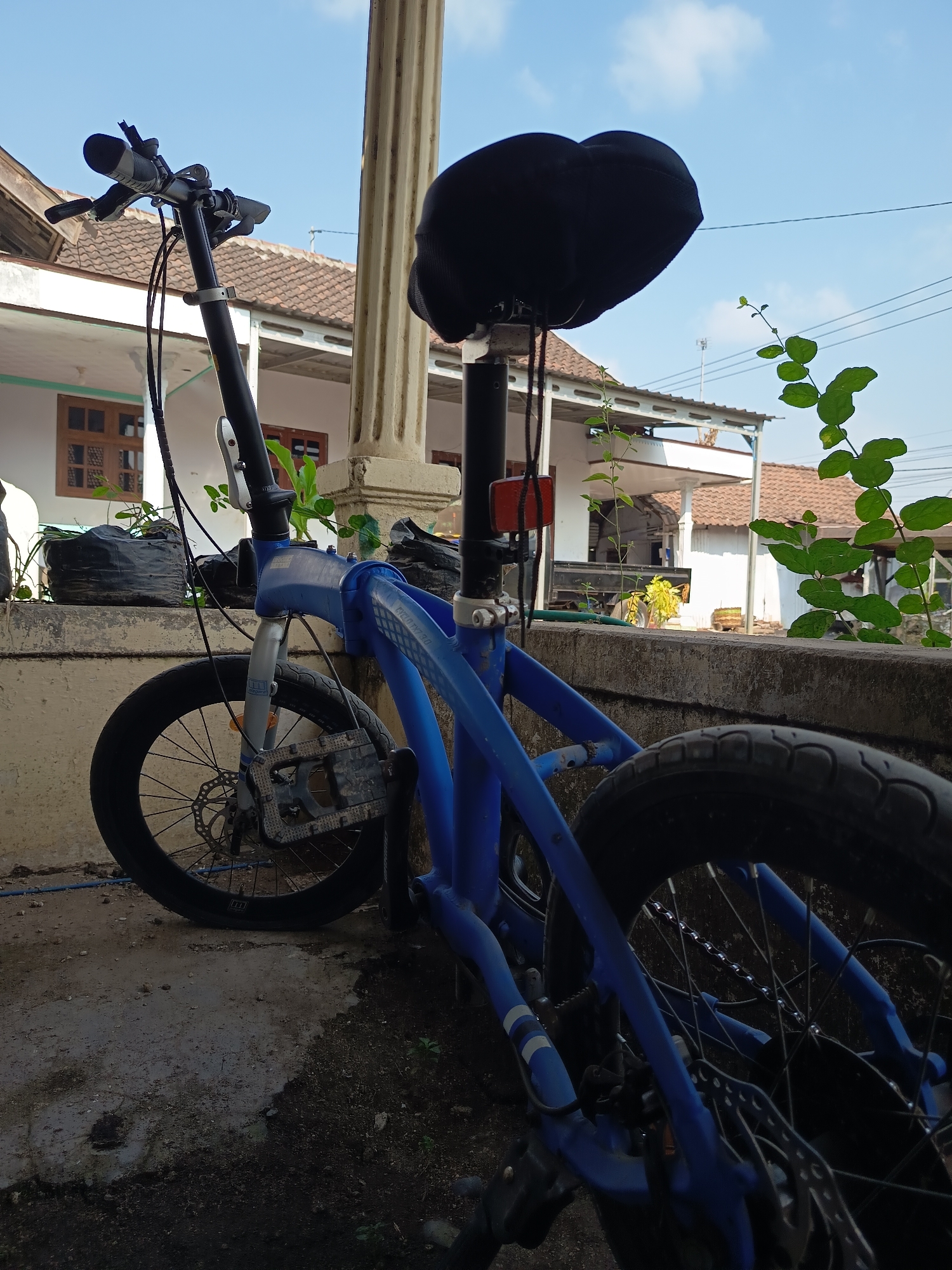 A blue folding bicycle is parked outdoors in what appears to be a residential area in Krampilan, Indonesia. The bicycle is positioned on a concrete surface with some dirt and vegetation around it. In the background, there is a house with a tiled roof and a concrete wall in the foreground. A large, dark bicycle seat is prominently visible. The sky is blue with some clouds. The scene suggests a casual outdoor setting, possibly a yard or driveway.