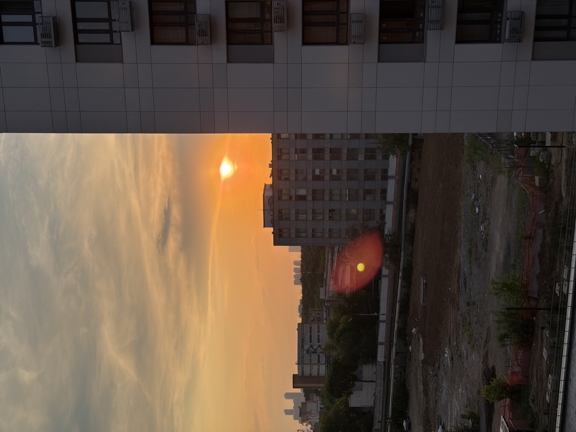 The image depicts a sunset over a city, likely Korolyov, Russia, featuring a modern apartment building in the foreground. The sky is ablaze with orange and yellow hues, with wispy clouds scattered across it. The sun, a bright orb, is visible slightly off-center, casting a warm glow. A distinct lens flare, appearing as a red and yellow circle, is present in the middle of the frame.
In the background, several buildings of varying architectural styles are visible. One prominent building, a multi-story structure with numerous windows and air conditioning units, dominates the middle ground. To the left, more distant buildings and what appear to be industrial structures can be seen against the horizon, some with a hazy silhouette due to the atmospheric conditions.
The foreground and middle ground reveal a construction or undeveloped site with disturbed earth, rubble, and some sparse vegetation. A fence, likely for construction site demarcation, is also visible. The overall scene is one of urban development juxtaposed with the natural beauty of a sunset. No people are clearly discernible in the image.