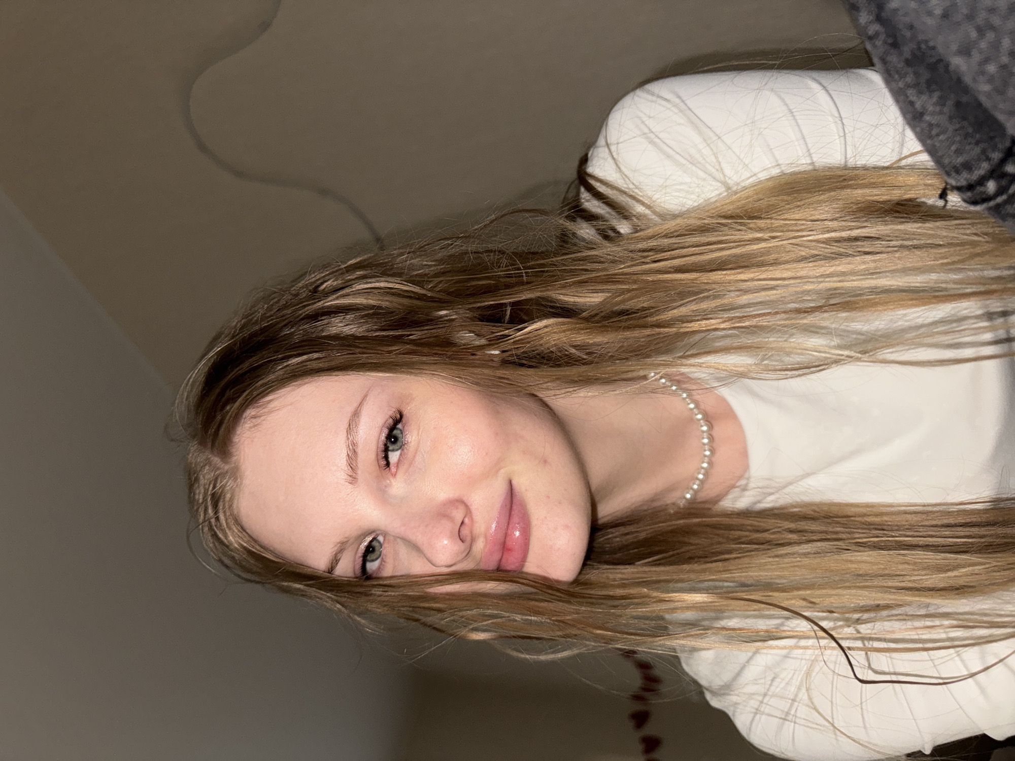 The media file features a young woman posing for a close-up, rotated portrait. She has long, wavy hair with shades of light brown and blonde, which is spread out across her white top and the plain background. Her head is positioned towards the left of the frame. She wears a simple white top and a pearl necklace. Her face, seen from a slight angle, displays light-colored eyes (appearing blue or green) and a gentle, pleasant smile, with soft pink lip gloss evident. The background is a plain, light beige or brown wall, with a subtle wavy pattern visible in the upper left corner. The lighting appears to be soft and artificial, suggesting an indoor setting. There is a dark gray fabric visible in the upper right, possibly part of another garment or bedding. No text is visible in the image. The image is taken in Perm, Russia.