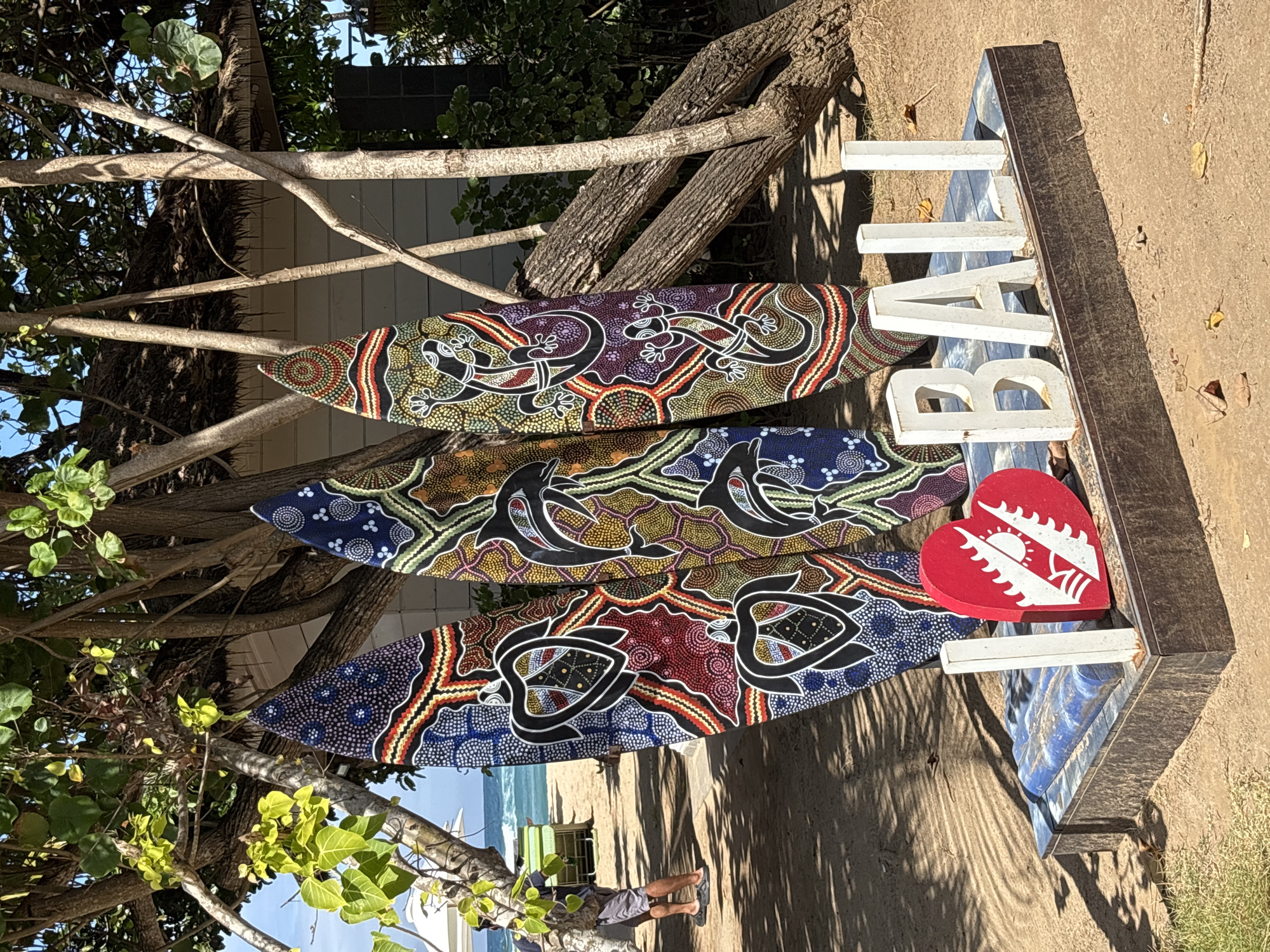 Three vibrantly decorated surfboards stand upright on a beach in Kuta Selatan, Indonesia. The surfboards feature Aboriginal-inspired dot art in a variety of colors and patterns, with one depicting turtles, another dolphins, and the third lizards. In front of the surfboards, a decorative sign spells out "I BALI" with white block letters and a red heart-shaped emblem. The scene is set on a sandy beach with lush green trees and foliage in the background. A small building with a thatched roof is visible behind the trees. In the distance, the ocean can be seen, with a hint of a beach umbrella and chairs. A person wearing shorts and a backpack is partially visible on the left side of the image, walking away from the camera. The overall setting suggests a tourist-friendly beach location.