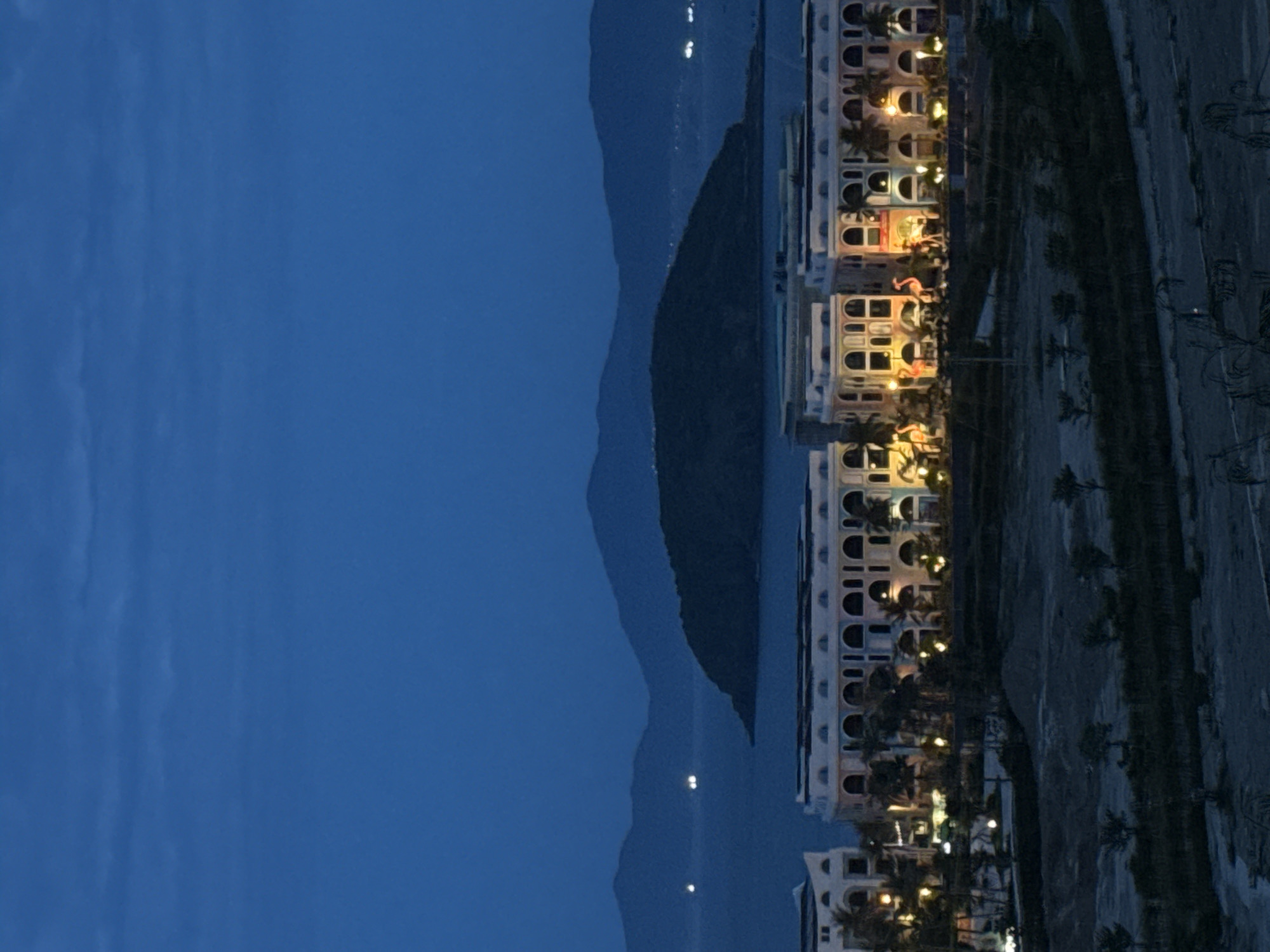 This is a long-shot, low-light photograph of a coastal scene at dusk or night in Bac Nha Trang Ward, Vietnam. In the foreground, a section of a modern, multi-story building complex with many arched windows and balconies is visible. The building is illuminated by warm lights, highlighting its facade. In front of the building, there are several large, decorative pink flamingo figures. Palm trees are also scattered throughout the area in front of the building.
Beyond the buildings, a body of water, likely the sea, stretches towards a dark, mountainous island in the middle ground. Further in the background, more distant mountains are silhouetted against the deep blue sky. There are several small, bright lights visible on the water, suggesting boats or distant coastal settlements. The overall atmosphere is calm and serene, with the artificial lights of the buildings contrasting with the natural darkness of the landscape. The ground in the immediate foreground appears to be rough and undeveloped, with some sparse vegetation.