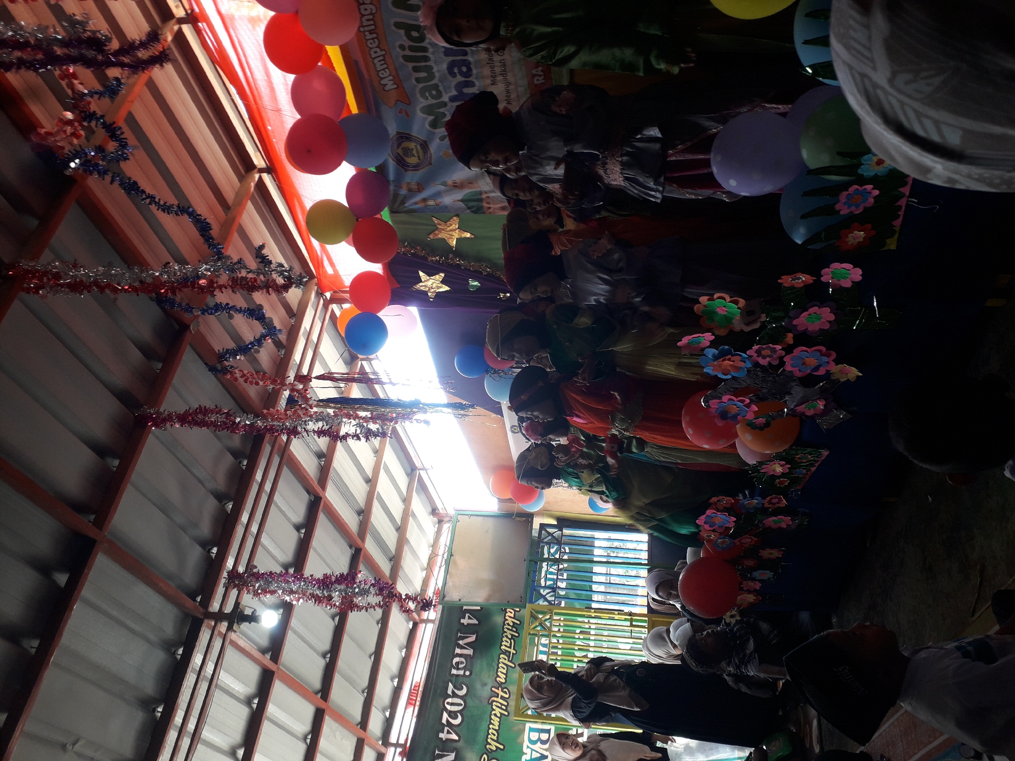 The image depicts an indoor event, likely a school or community gathering, held under a corrugated metal roof supported by a wooden structure. Red, white, and blue streamers hang from the ceiling, alongside bright red and white tinsel. Colorful balloons, including red, orange, yellow, blue, and purple, are arranged around the scene. A banner with Arabic script, possibly congratulating an event, is visible in the background, along with a smaller banner in Indonesian that reads "14 Mei 2024 N".
The foreground and middle ground are populated by a group of children and adults, many of whom are dressed in traditional Islamic attire. Several children in the middle ground appear to be performing or watching a performance on a raised platform. They are dressed in colorful robes and head coverings. Some of the children are holding small decorative items or have their hands raised. Adults in the background are also observing the event, with one woman in the left foreground holding up a mobile phone, possibly recording. The overall atmosphere suggests a celebratory occasion. The provided location of Cankuang, Indonesia, indicates a cultural context for the event.