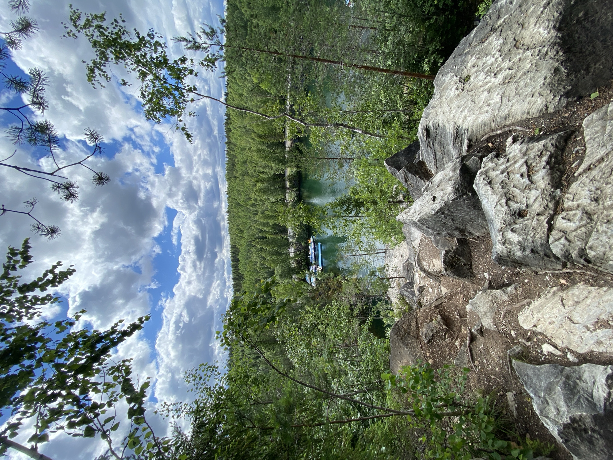 This is a high-angle shot of a rocky, wooded area overlooking a body of water. In the foreground, large grey rocks are scattered across the ground, with some vegetation growing between them. The path appears to be unpaved and leads down towards the water.
In the middle ground, the water is a deep green color, and several small boats are docked along the shore. The boats are light blue and white. Surrounding the water is a dense forest of green trees that extends to the horizon.
The sky above is a mixture of blue sky and white, fluffy clouds. The overall impression is one of a natural, serene landscape.
This scene is likely located in the Ruskeala Mountain Park in Ruskeala, Russia, a popular tourist destination known for its marble quarries and scenic beauty. The park features a large, flooded marble quarry that has been transformed into a picturesque lake.