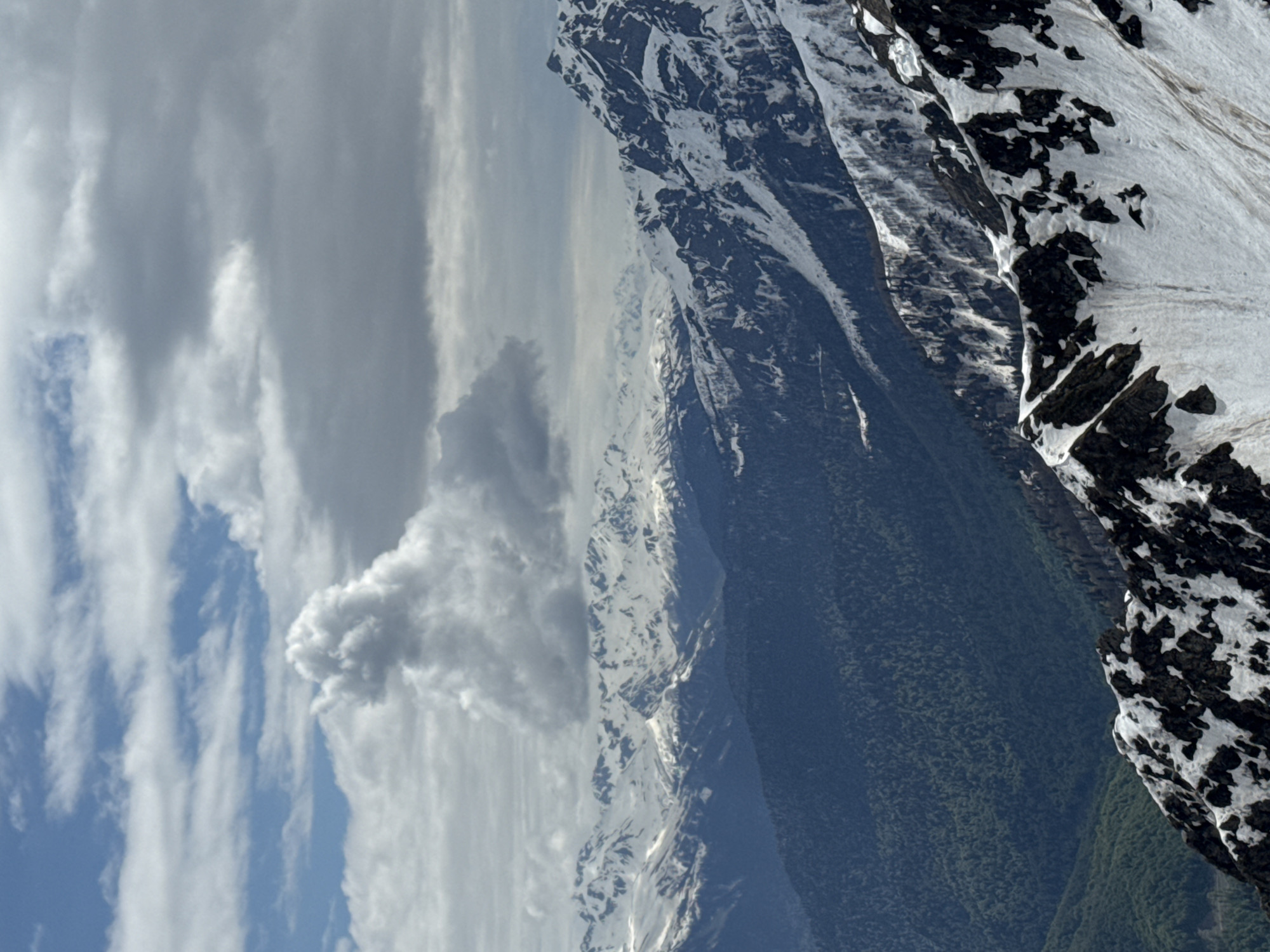 The image depicts a mountain landscape in Estosadok, Russia. Snow-capped peaks are visible, with some areas showing exposed rock and vegetation. A forest covers the lower slopes. The sky contains a mix of blue and gray clouds.
