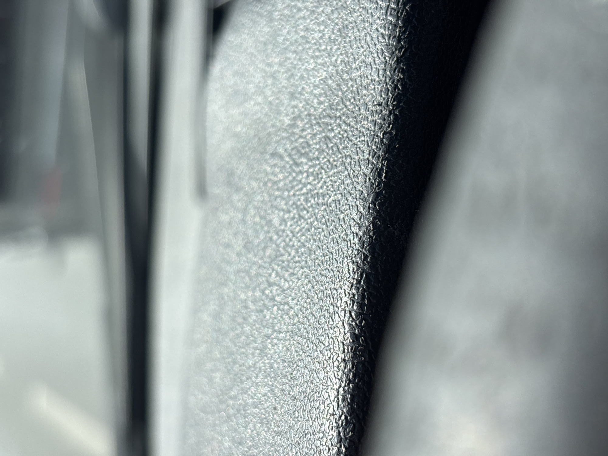 The image depicts a close-up, low-angle shot of a textured, dark gray surface, likely a dashboard in a vehicle. The texture appears to be a fine, granular pattern, characteristic of many automotive interiors. The lighting suggests an indoor environment, possibly with ambient light reflecting off the surface. The background is out of focus, showing blurred shapes and colors that could indicate the interior of a car, such as a windshield or other dashboard elements. There are no people or discernible landmarks visible that would allow for specific identification of a location within Minsk, Belarus. The scene is devoid of any action or interaction.