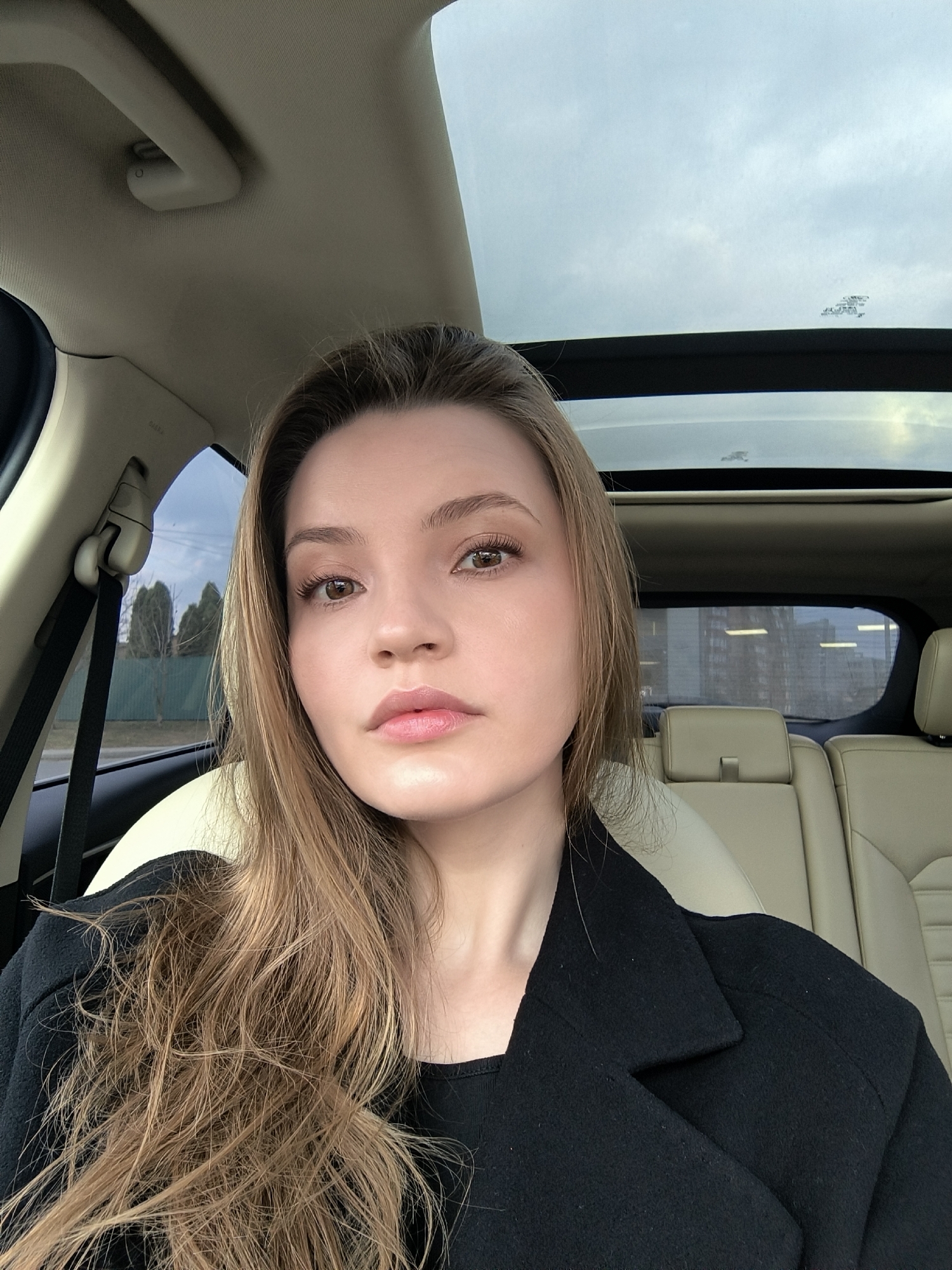 A young adult female is depicted in a selfie, seated in the front passenger seat of a vehicle in Vinnytsia, Ukraine. She has long, light brown hair, brown eyes, and fair skin, with visible light makeup. She is wearing a black long-sleeved top beneath a black overcoat or blazer. Her gaze is directed towards the camera. The vehicle interior features light beige leather seating, including the headrests for the front passenger and rear seats. A panoramic glass sunroof reveals an overcast sky. A beige grab handle and an "AIRBAG" label are visible on the vehicle's interior roof lining. Through the open side window to the left, an asphalt road, a green metal fence, and deciduous trees with bare branches are visible. Through the rear window, distant multi-story residential buildings are discernible. The immediate environment suggests a roadside or residential area within a city.