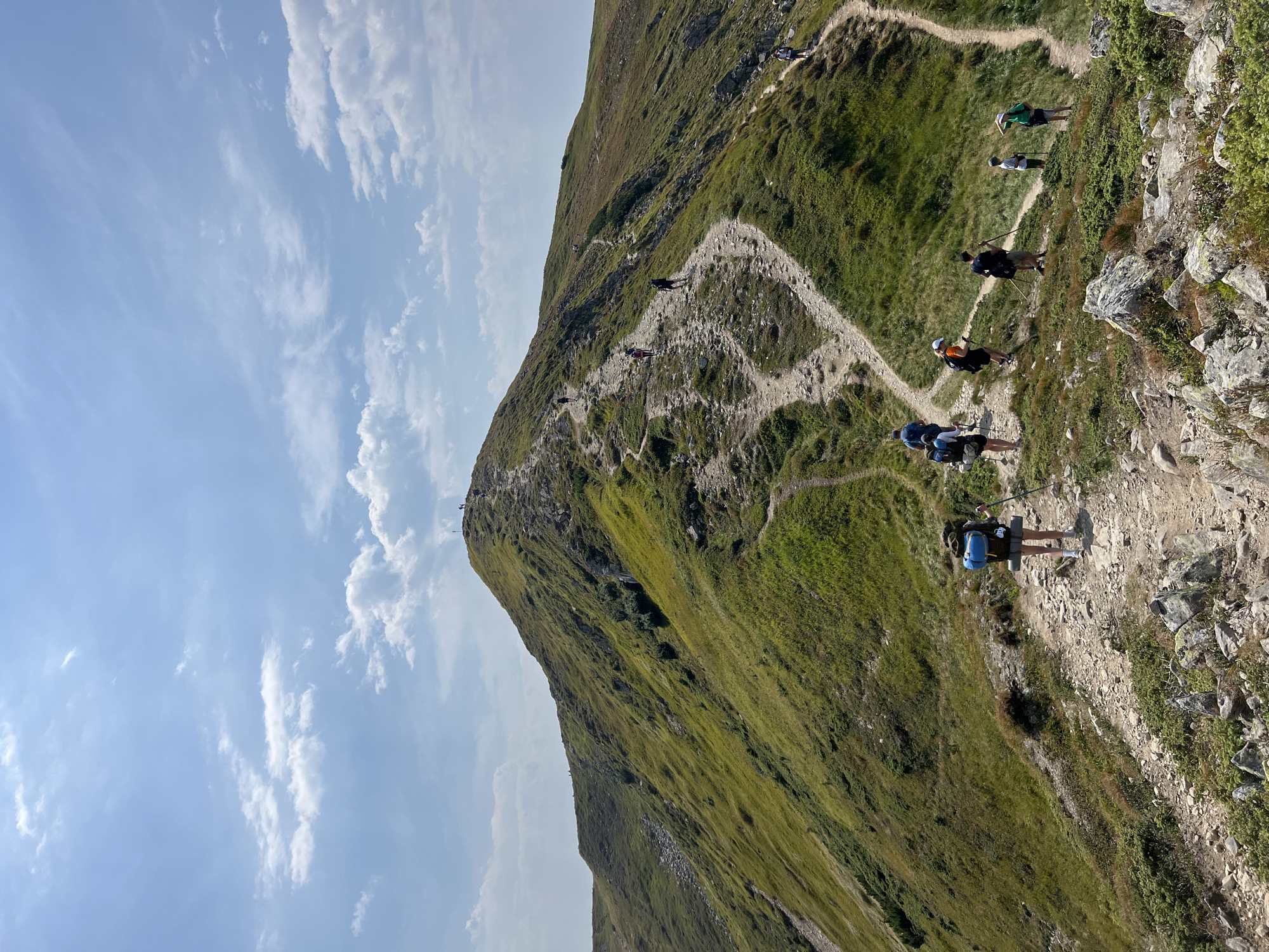 A group of hikers are ascending a steep, grassy mountain path under a bright blue sky with scattered white clouds. The path is narrow and rocky, winding its way up the side of a verdant mountain. The hikers, some wearing backpacks and using trekking poles, are spaced out along the trail, moving towards the summit. The landscape is characterized by rolling green hills and rocky outcrops. The image captures a scenic view of the Carpathian Mountains near Vorokhta, Ukraine, a popular destination for outdoor activities.