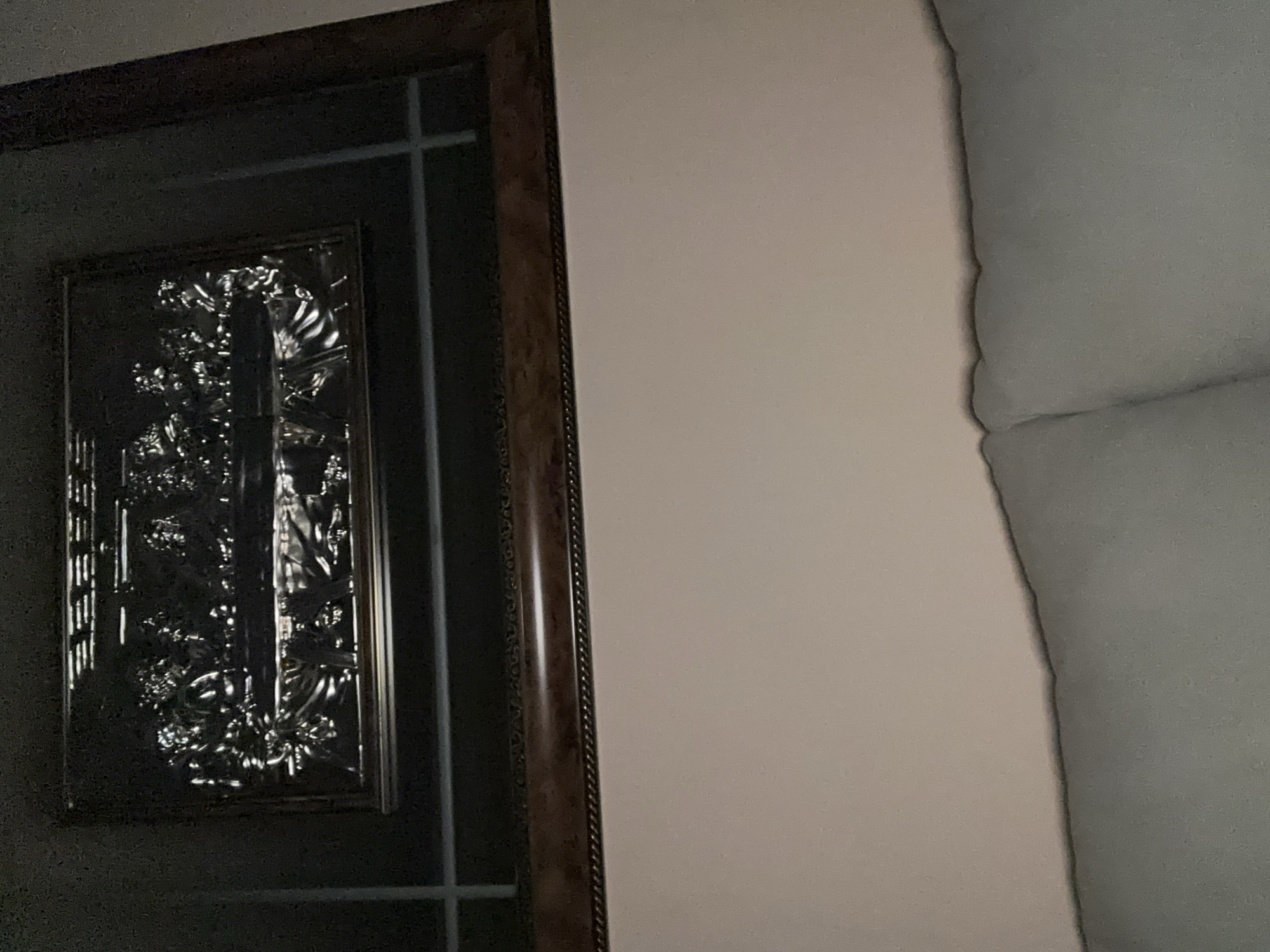 A dark wooden framed object, possibly a mirror or decorative panel, hangs on a light beige wall. The framed object features a metal insert with ornate floral and geometric patterns. To the right of the wall, part of a light gray object or structure is visible. No specific location in Yerevan, Armenia, can be determined from the image. No people, actions, or interactions are visible.