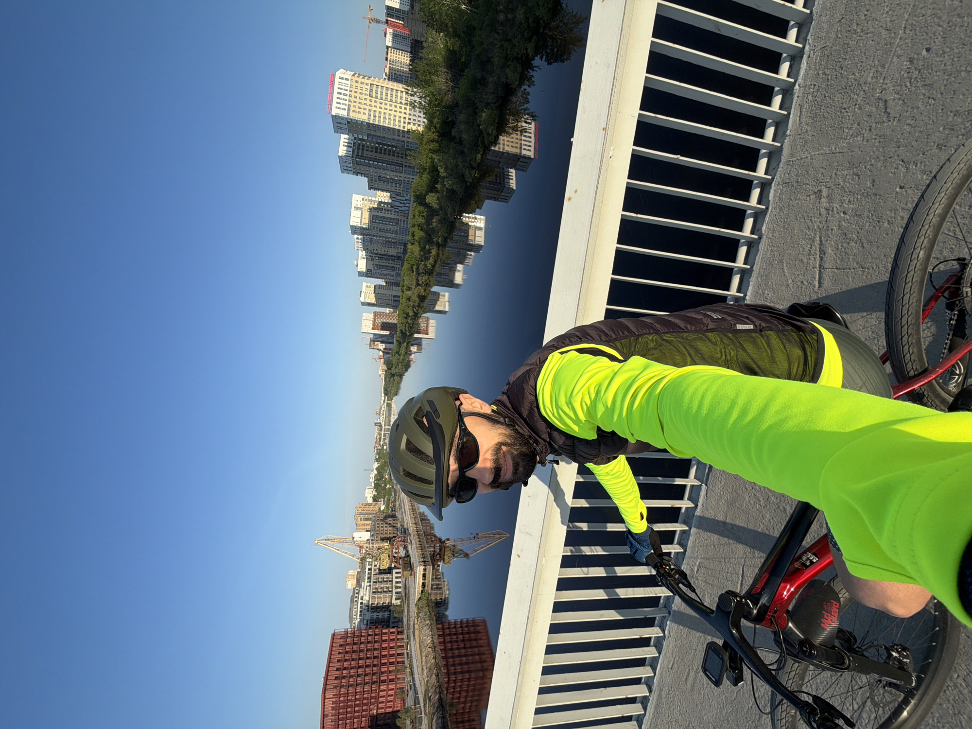 A man is riding a bicycle across a bridge in Tyumen, Russia. The man is wearing a helmet, sunglasses, and a bright neon yellow shirt and vest. He is holding the handlebars of the bike and looking toward the camera. The bridge has a white metal railing with black spaces in between. In the background are modern high-rise buildings, construction cranes, trees, and a blue river reflecting the buildings and trees. The sky is clear and blue. The scene suggests a sunny day in an urban setting in Tyumen, Russia.
