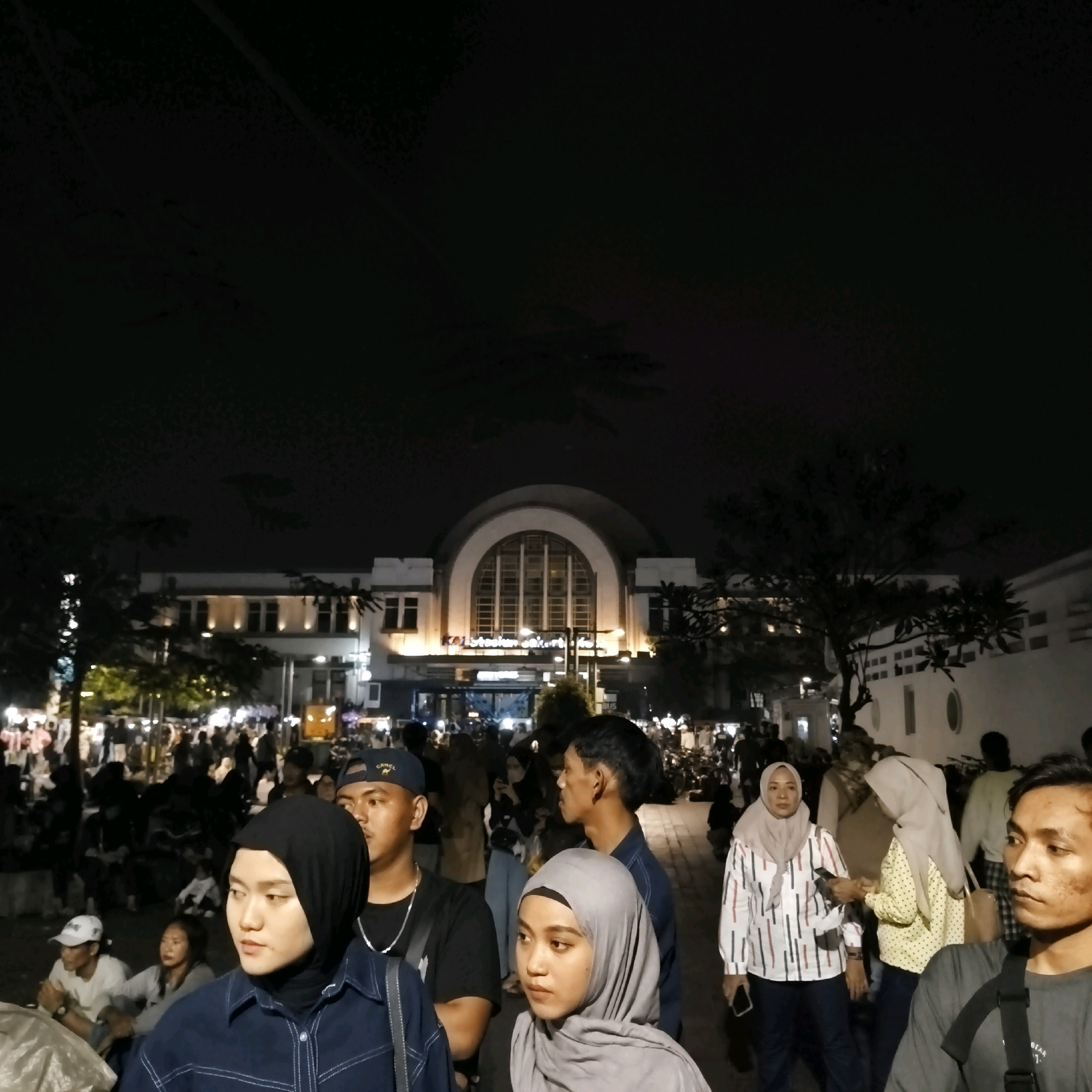 The image shows a nighttime crowd gathered in front of a large building with a prominent arched window, identified as the historic Jakarta Kota railway station (Stasiun Jakarta Kota) in West Jakarta, Indonesia. The building is illuminated, highlighting its architecture. Many people are visible, some in the foreground and many more in the background, suggesting a public event or gathering. Several individuals are wearing hijabs, common attire for Muslim women in Indonesia. The atmosphere appears lively, with the presence of many people and the illuminated building. The lighting suggests it is evening or night.