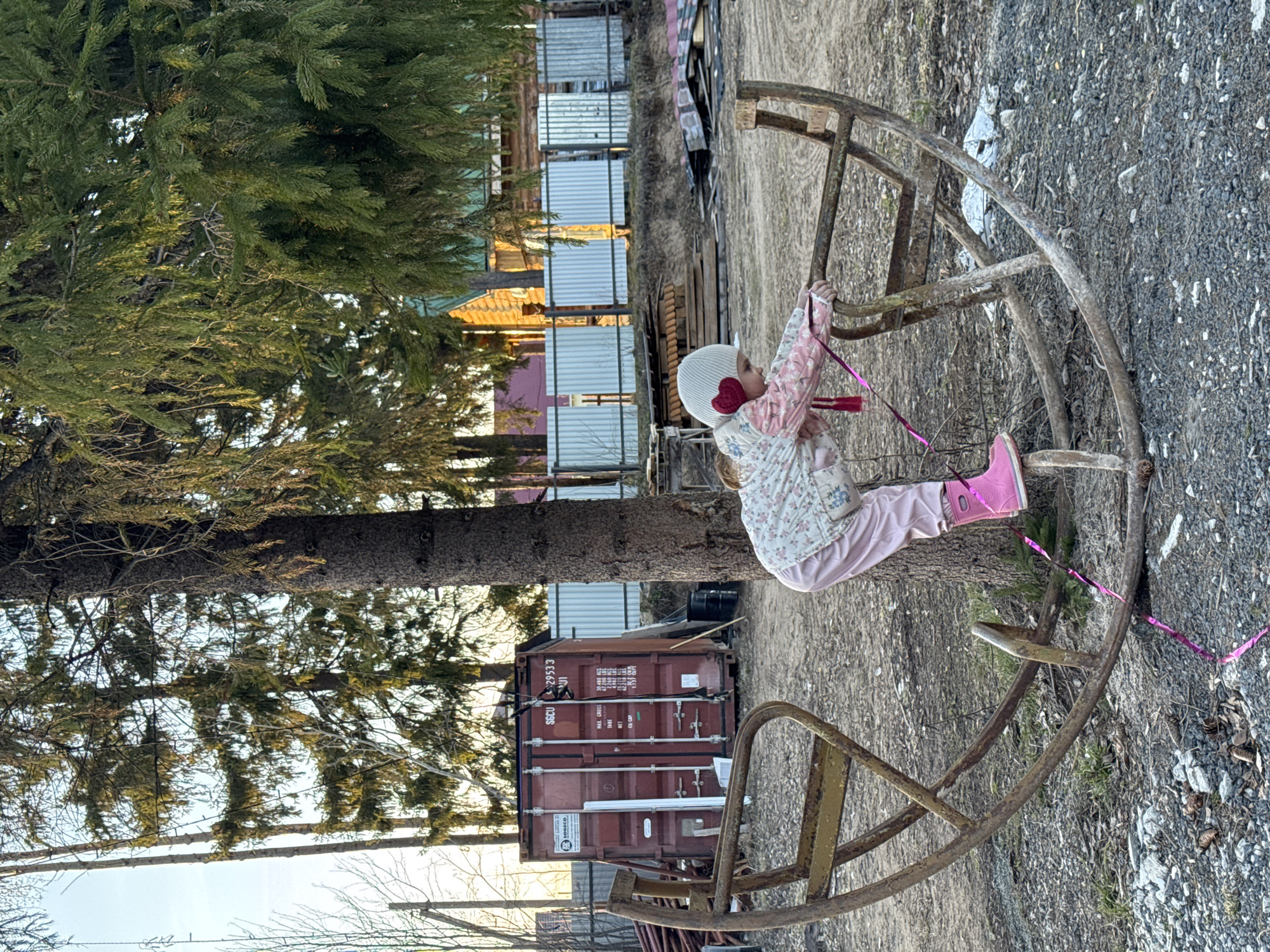 A young child is playing on a rusty, circular metal structure in an outdoor environment. The child is wearing a white hat with red heart-shaped embellishments, a floral patterned jacket, light pink pants, and bright pink boots. The child holds onto the metal structure with both hands. A shiny pink ribbon is tied to one section of the metal.
The background includes a tall evergreen tree on the left. Behind the tree, a brown shipping container is visible, and further back, there are buildings with a light-colored fence in front of them. The ground is covered in gravel and small vegetation.
The location is identified as Chaikovsky, Russia.