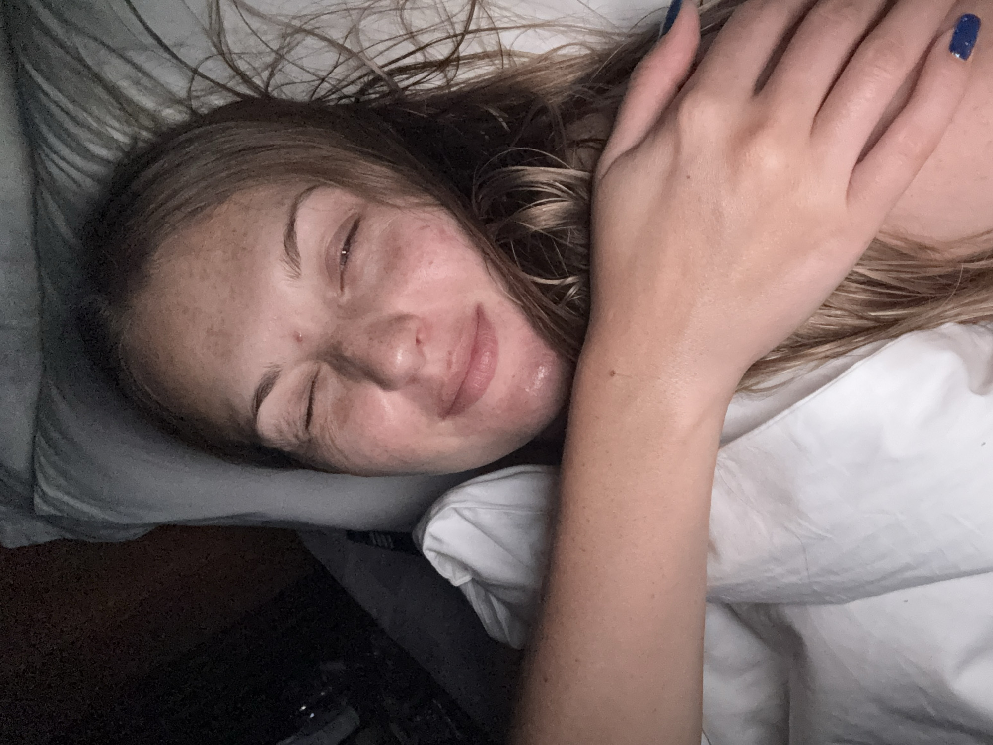 A woman is lying in bed, her face partially obscured by her arm. She has fair skin with freckles and light brown hair that is spread around her head. Her eyes are closed, and she is smiling slightly. She is wearing a white top. Her left arm is bent and resting across her face, with her hand covering her forehead and right eye. Her fingernails are painted a dark blue. The bed appears to have white sheets and a grey pillow. The background is dark and appears to be a bedroom. There is no indication of the city of Denpasar, Indonesia in the image.
