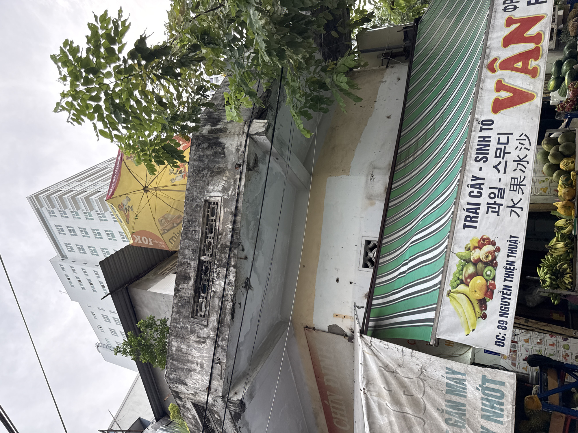 The image shows an outdoor scene in Nha Trang, Vietnam, focused on a fruit stand below a dilapidated building. A large, white, multi-story modern building dominates the background, with a cloudy sky overhead. In the mid-ground, an older, weathered concrete structure with a sloping metal roof is visible, adorned with a yellow umbrella featuring text and graphics. A green and white striped awning extends from this building, partially obscuring the fruit stand below. The fruit stand displays a variety of fresh fruits, including bananas, grapes, pears, strawberries, kiwis, and melons, as well as signage with Vietnamese text and a prominent red "VAN" logo. The overall impression is of a busy urban environment with a mix of old and new architecture, featuring a vibrant local fruit market.