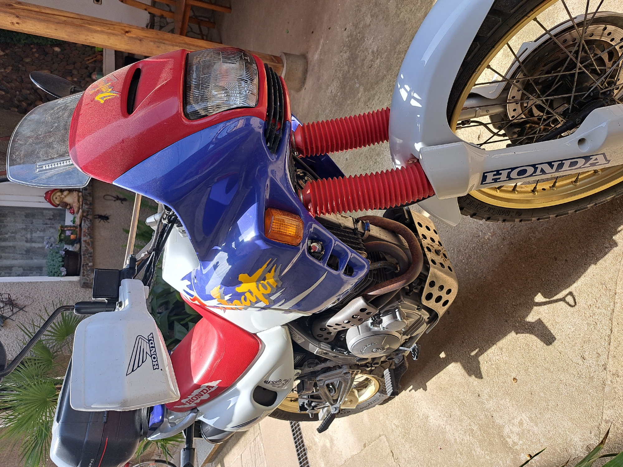 The image shows a Honda motorcycle parked on a concrete surface. The motorcycle is red, white, and blue. The front wheel is visible, and the tire has gold rims. The motorcycle has a windshield and red fork gaiters. The background is blurred, but includes a glimpse of a building, plants, and wooden structures. The location is likely Chepintsi, Bulgaria, though that cannot be confirmed from the image alone.