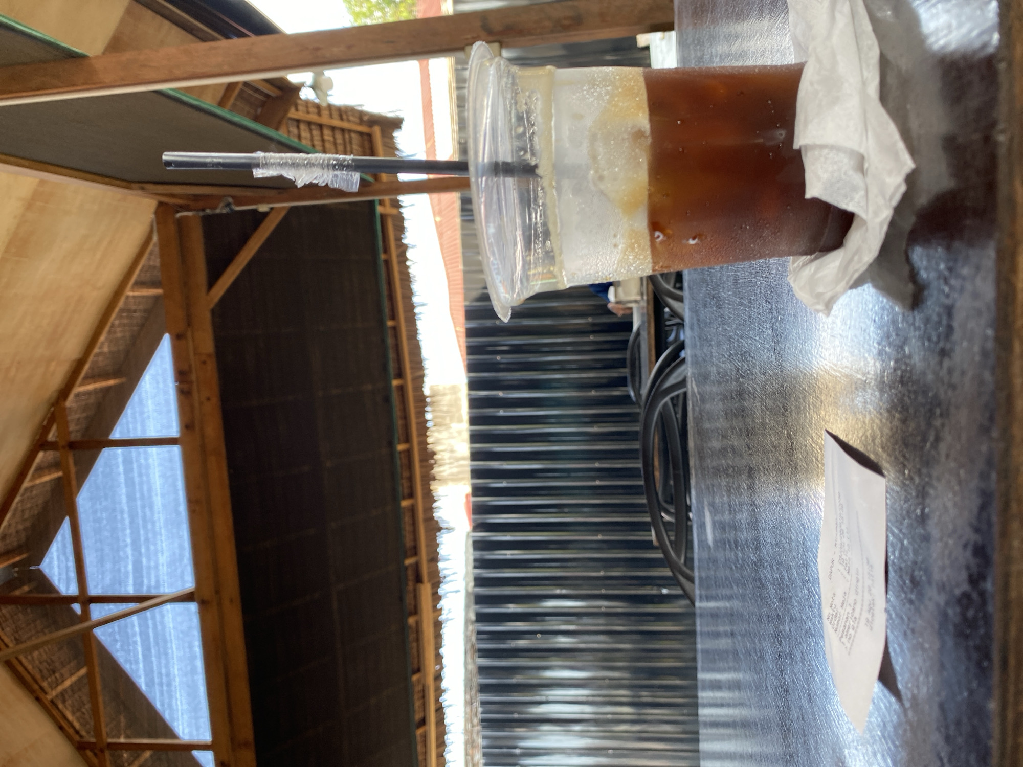An iced coffee sits on a dark, reflective table in the foreground. The cup is clear plastic, filled with a dark liquid and ice, with a black straw extending from the top. A white receipt is partially visible on the table to the left of the cup, and a crumpled white napkin is beneath the cup. Behind the table, a corrugated metal fence runs horizontally, and beyond that, rooftops and trees are visible. The scene is set under a thatched roof structure with visible wooden beams and supports, suggesting an outdoor or semi-outdoor dining area. The lighting indicates it is daytime. The image likely depicts a cafe or restaurant in Lhokseumawe, Indonesia, given the style of the architecture and the Indonesian text on the receipt. No individuals are clearly visible in the frame, though blurred figures and chairs suggest a public space.