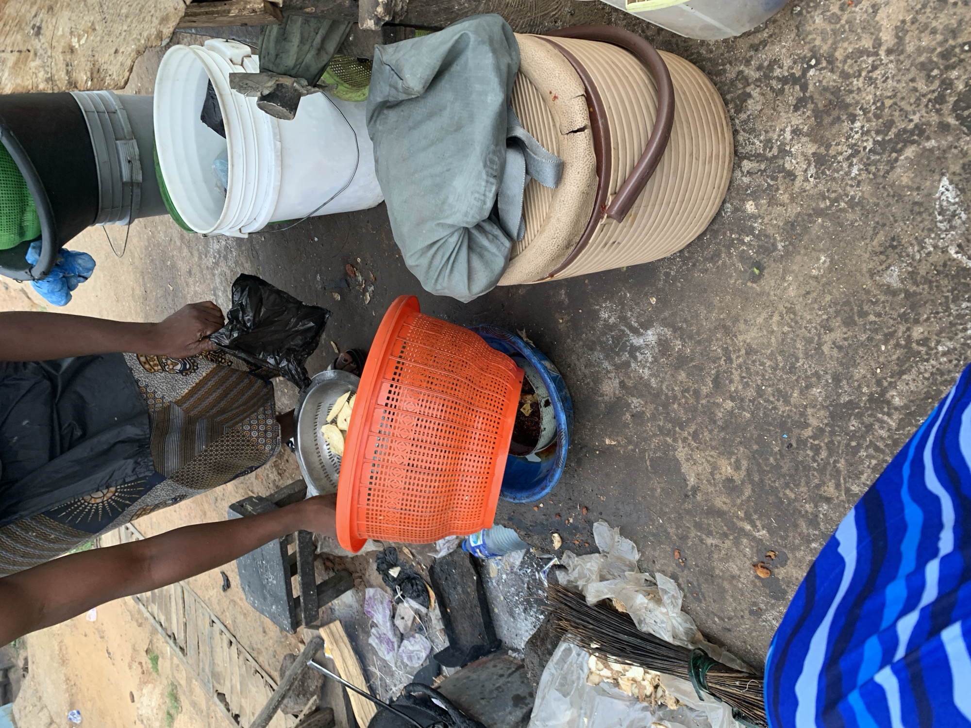 In New Stadium, Nigeria, an outdoor scene displays multiple objects on a soiled, concrete surface. At least one person is visible, evidenced by dark-skinned arms and hands manipulating a black plastic bag near a silver strainer containing food items. An orange plastic basket rests at an angle, appearing to be near a blue plastic container. Several buckets of varying colors (black, white, and gray) are stacked adjacent to a light brown, lidded container draped with gray fabric. Discarded plastic and wooden debris are scattered around the lower portion of the frame. A bundle of dark sticks, bound with green material, lies near the right edge of the frame, next to an area with blue striped fabric.