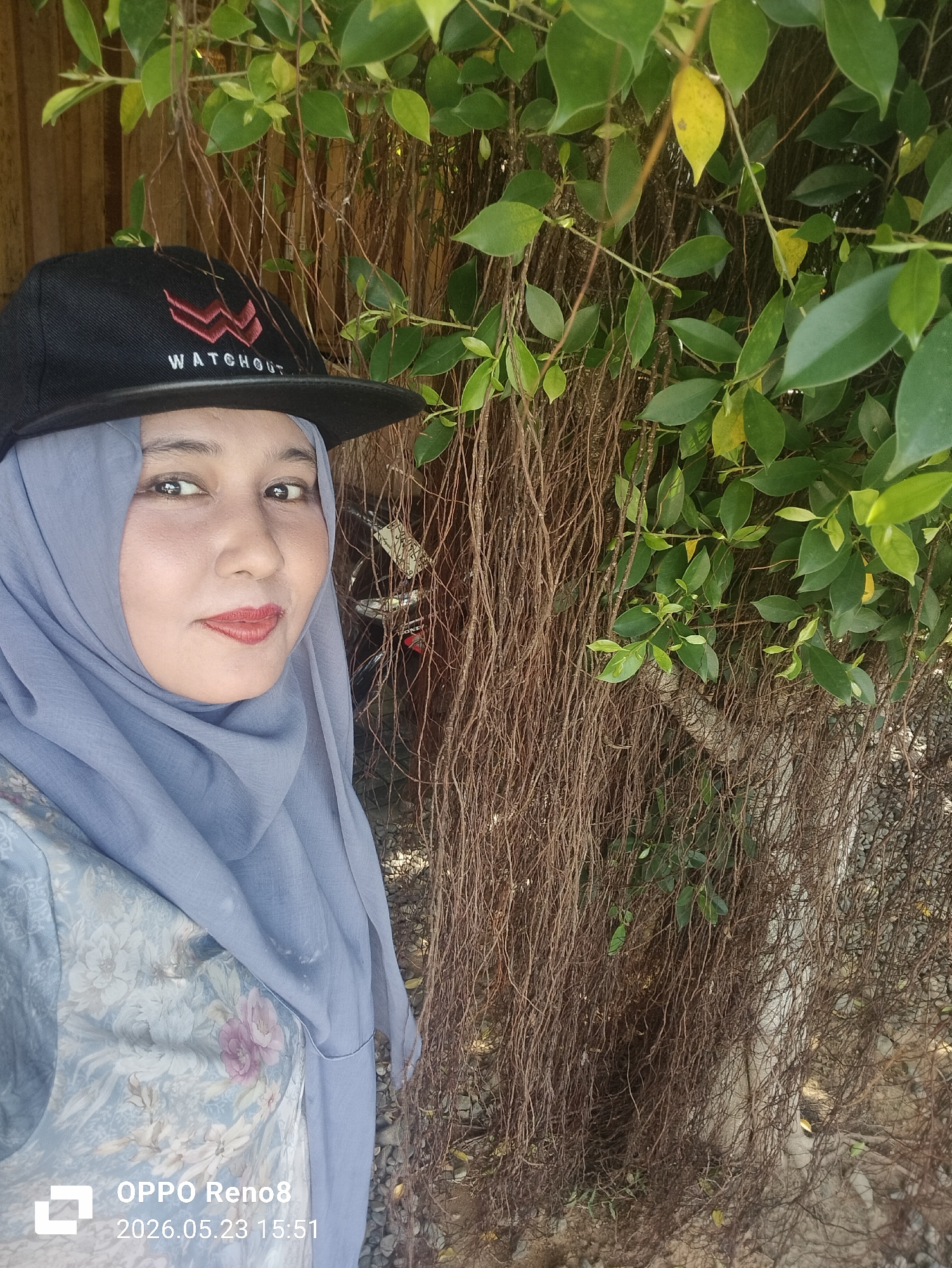 A woman wearing a blue hijab and a black baseball cap stands facing the camera. The cap has the word "WATCHOUT" on it. The woman is wearing a floral patterned garment. She appears to be standing in front of a wall of vines and leaves. The lower left corner of the image includes the text "OPPO Reno8 2026.05.23 15:51". The location is implied to be Santan, Indonesia.
