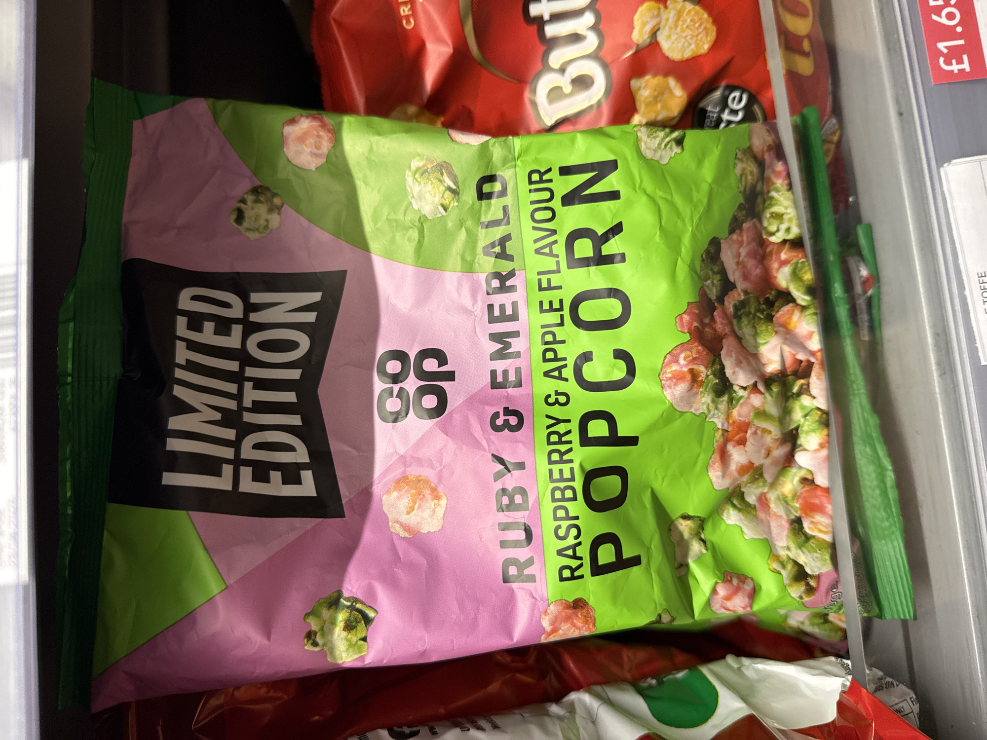 The image shows a package of Co-op Limited Edition Ruby & Emerald Raspberry & Apple Flavour Popcorn, positioned in what appears to be a refrigerated shelf or display case. Other food packages are partially visible in the background, including a package of "Butter" flavored snacks and a label indicating a price of "£1.65" for "TOFFE". The overall environment suggests a retail setting, possibly a grocery store.