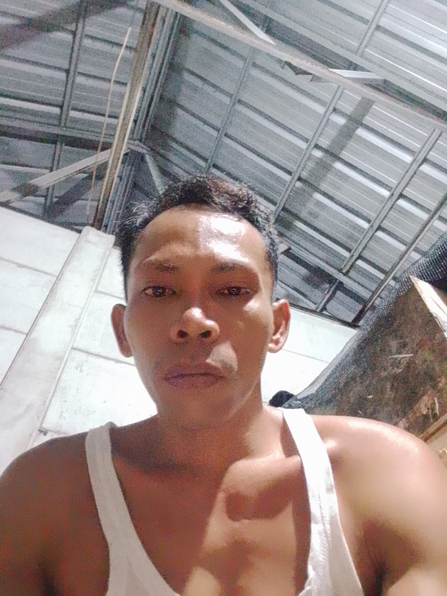 A close-up shot of a man with short dark hair, wearing a white tank top, facing the camera. His skin appears to have a sheen of moisture. The background shows a corrugated metal ceiling supported by a framework of metal beams. The location is possibly Bukit Ketok, Indonesia. There are hints of a white wall to the left and a darkened, possibly cluttered, area to the right.