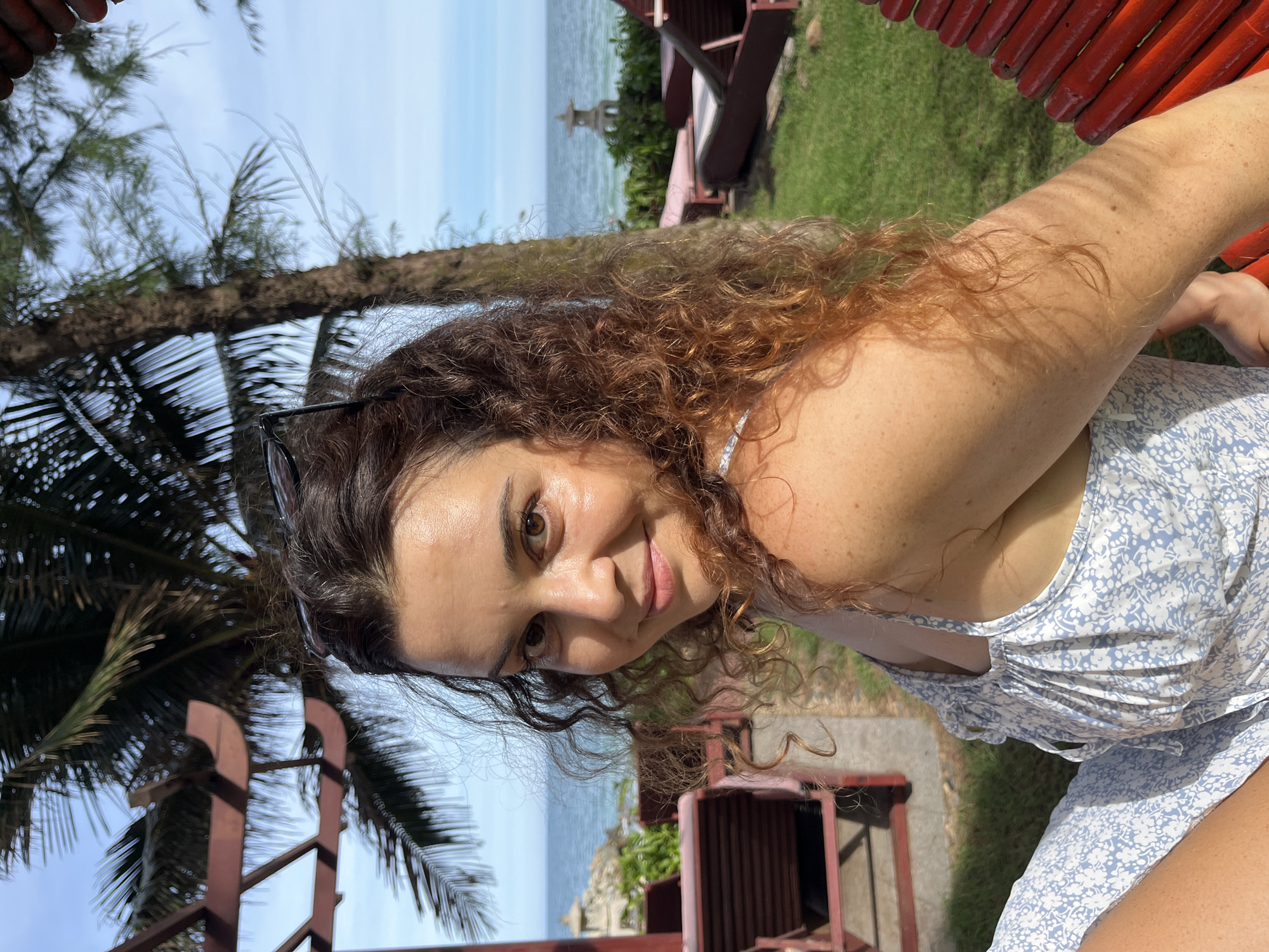 A medium shot captures a woman with curly brown hair and fair skin sitting outdoors in Mũi Né, Vietnam. She is wearing a light blue and white floral dress and has sunglasses perched on her head. The woman is looking towards the camera with a slight smile. In the background, a palm tree and the ocean are visible, along with lounge chairs and greenery, suggesting a resort or beachfront location. The lighting indicates it is daytime, with sunlight casting shadows.