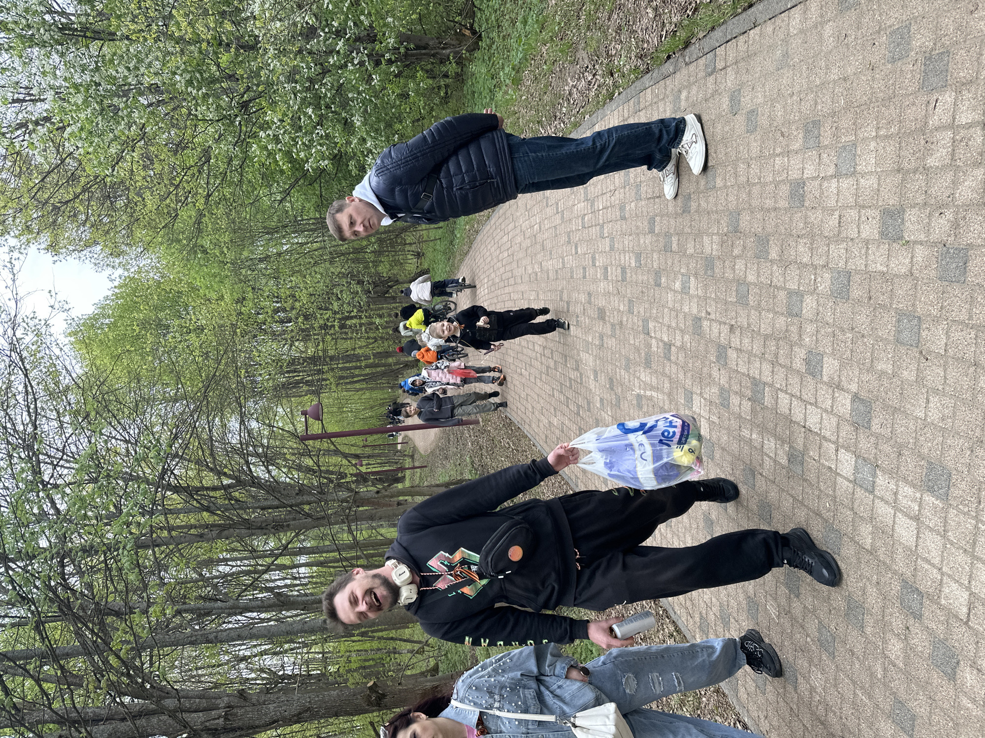 The scene shows a paved path in Grebnevo, Russia, lined with trees. A group of people are walking along the path. In the foreground, a man is holding a plastic bag and a can. Further down the path, people are walking and cycling. Another man is standing with his arms crossed. The trees are in bloom, suggesting it is springtime.