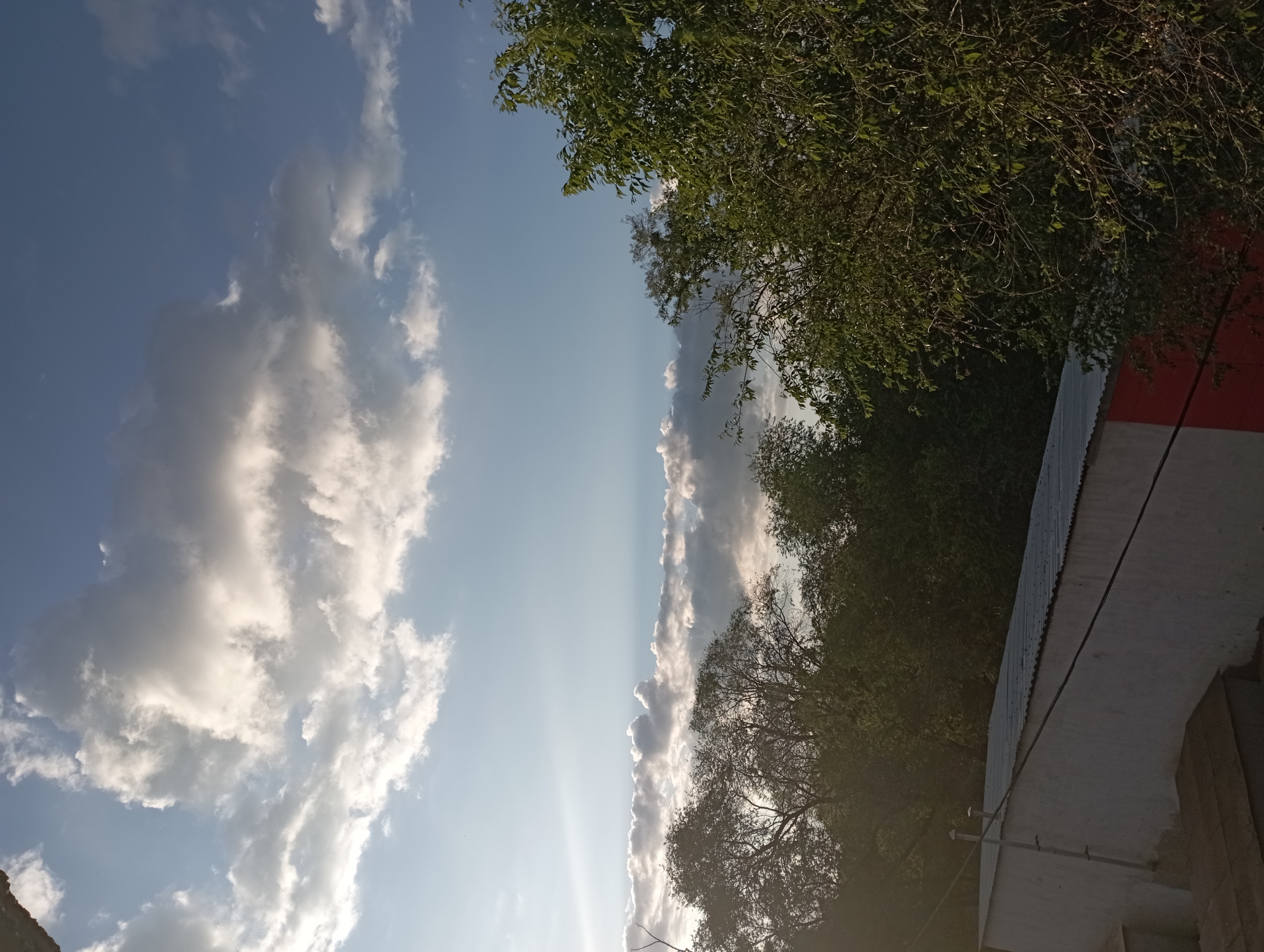 The image depicts a view of the sky with clouds, trees, and a building with a corrugated metal roof. The sky is a mix of blue and white, with large, fluffy clouds dominating the upper portion and a band of darker clouds near the horizon. Sunlight is visible, casting a bright glow and creating lens flare on the left side of the image. A line of green trees with dense foliage forms a boundary between the sky and the lower part of the scene. In the foreground, a building with white walls and a red section is partially visible, topped by a gray corrugated metal roof. The overall impression is of an outdoor scene, possibly in a semi-urban or suburban area, during daylight hours. The specific location in Blagoveshchensk, Russia, cannot be determined from the visual information provided. There are no people or specific actions captured in the image.