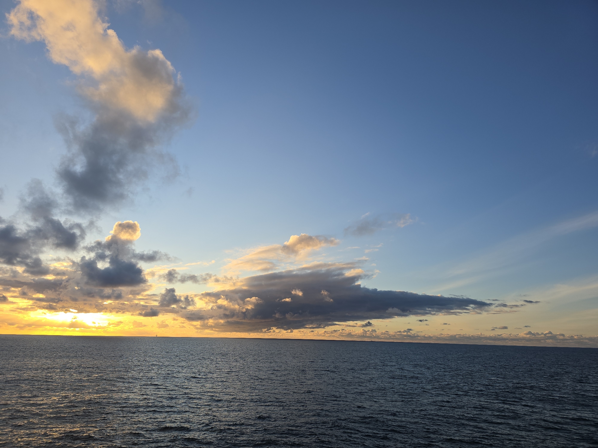 The image depicts a seascape at sunset. The sky is predominantly blue, transitioning to orange and yellow near the horizon. Clouds of varying shapes and sizes are scattered across the sky, some reflecting the colors of the sunset. The sea is dark blue with subtle ripples. The horizon line is visible, with a strip of brighter light indicating the setting sun. The location is unknown.