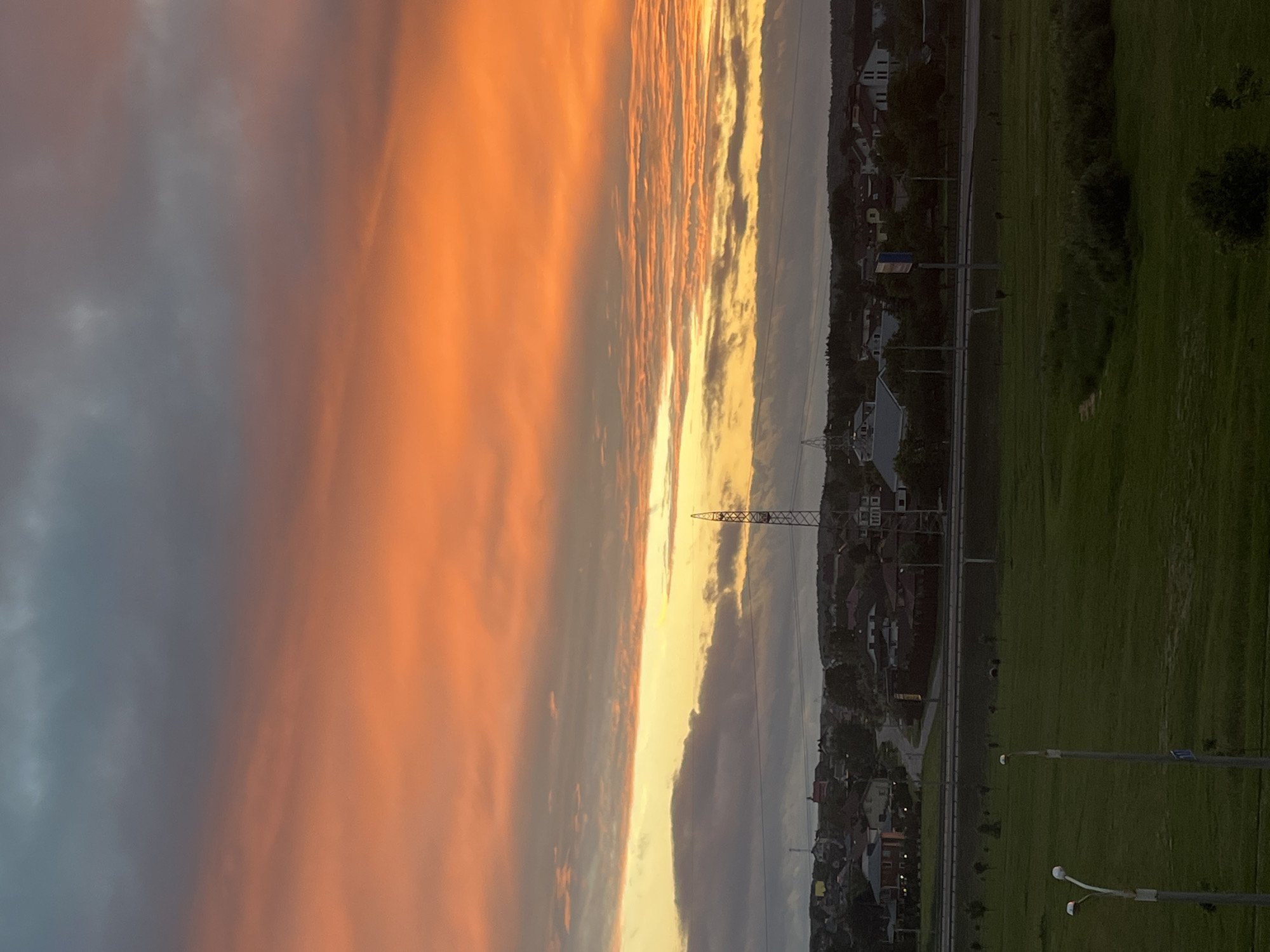 An aerial view captures a vibrant sunset over a suburban landscape in Minsk, Belarus. The sky is dominated by a dramatic gradient of fiery oranges, pinks, and yellows, with streaks of dark gray clouds adding depth. Below the sky, a power line tower stands prominently, silhouetted against the colorful horizon. The middle ground reveals a residential area with houses of various sizes and architectural styles, interspersed with trees. A road or highway runs horizontally across the scene, separating the houses from a large, grassy field in the foreground. The field is dotted with sparse trees and features two streetlights with white globes. The overall impression is one of a peaceful evening in a developing urban area.