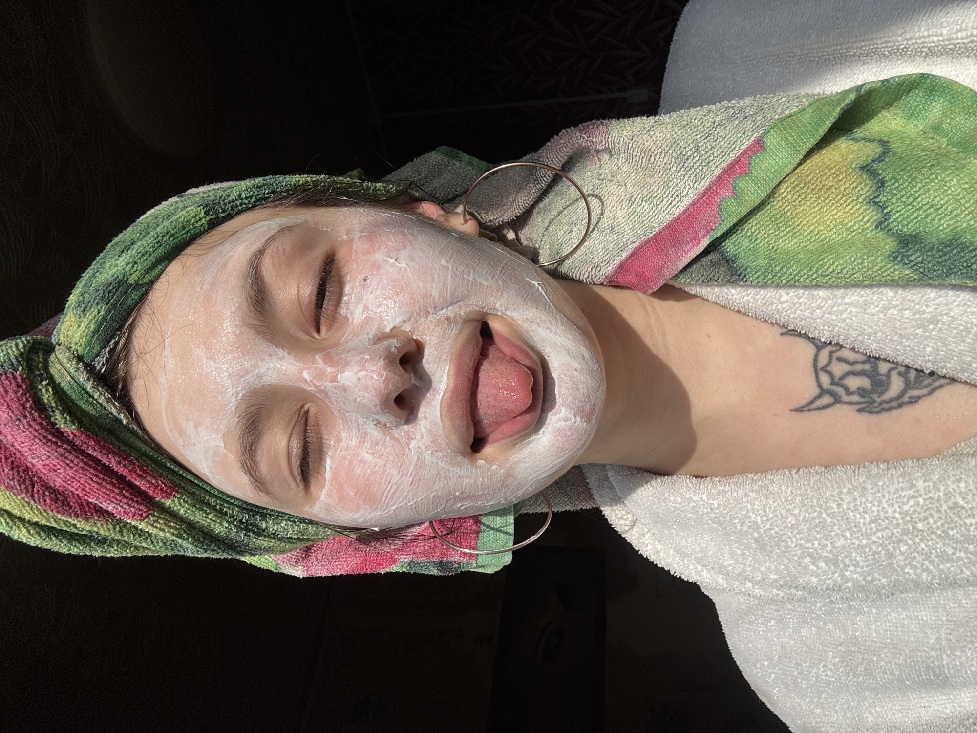 A person with closed eyes, covered in a white facial mask, is lying down, sticking out their tongue. The person wears a towel wrapped around their head, with green, pink, and white colors. A gold hoop earring is visible. The person is wearing a white robe. A tattoo is visible on the upper chest. The location is unknown.