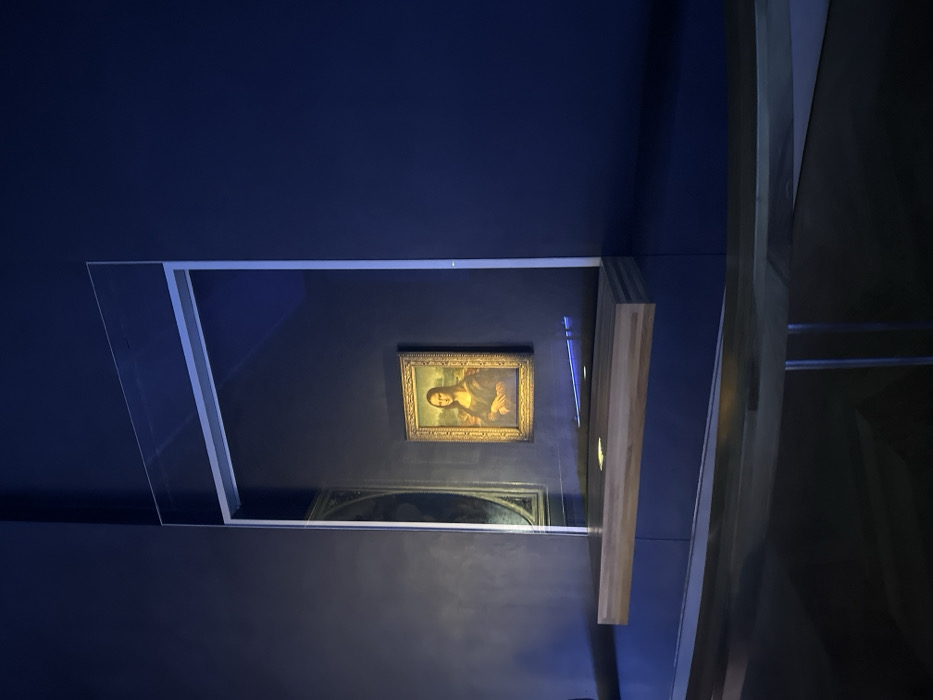 The image depicts the Mona Lisa painting by Leonardo da Vinci, located in the Louvre Museum in Paris, France. The painting, presented in an ornate gold frame, is centrally positioned on a deep blue wall within a dimly lit gallery. A large, transparent protective glass panel encloses the artwork, reflecting ambient light and partially obscuring another darker, circular artwork to its left. In front of the glass panel, a light-colored wooden ledge or barrier extends horizontally, seemingly suspended. The foreground reveals a dark, curved railing, indicating a viewing area for visitors. The scene is devoid of people, emphasizing the artwork and its protective enclosure, which is brightly illuminated against the dark background.